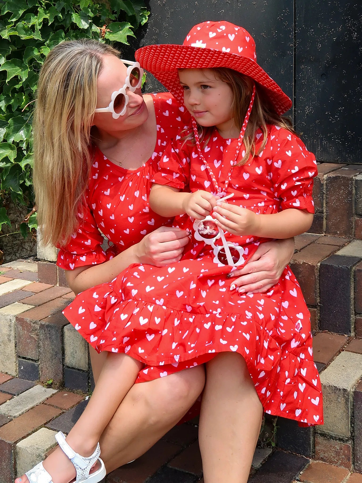 A woman and young girl sit on brick steps in matching Hearts Red & Pink Heart Midi Dresses. The girl accessorizes with a red hat and white sunglasses—adorably styled, ethically made, and perfect for Mum & Me looks.