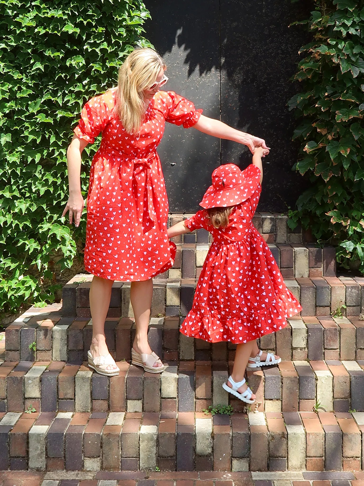 An adult and child in matching Hearts Red & Pink Heart Midi Dresses and sandals stand hand in hand on brick steps, with the child also wearing a matching sun hat. Ethically made by Hearts, with ivy framing both sides.