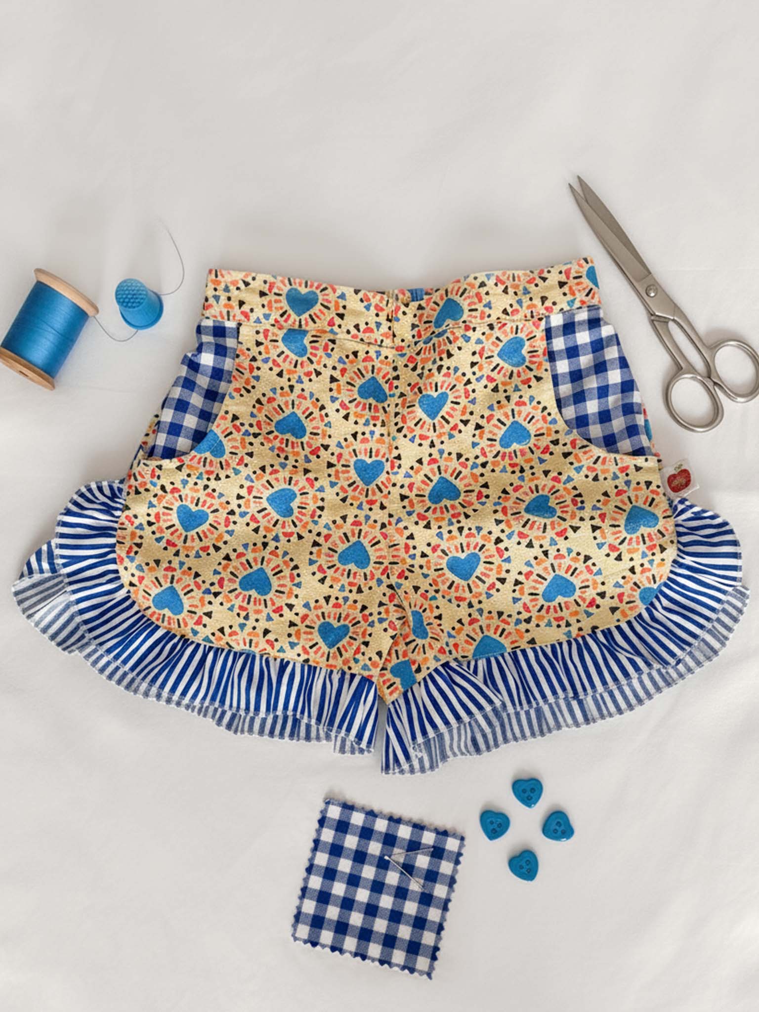 Starburst Ruffle Shorts by Starburst feature a yellow floral print, blue gingham accents, striped ruffle trim, sewing scissors, blue thread spool, blue buttons, and a matching gingham square. Ethically made in India.