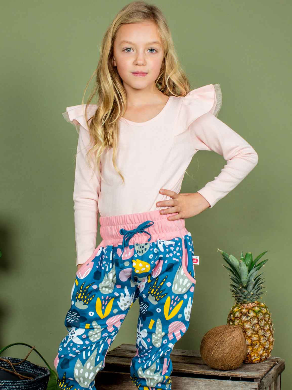 Young girl wearing a pink top and blue patterned pants standing against a green wall with plants and a pineapple.