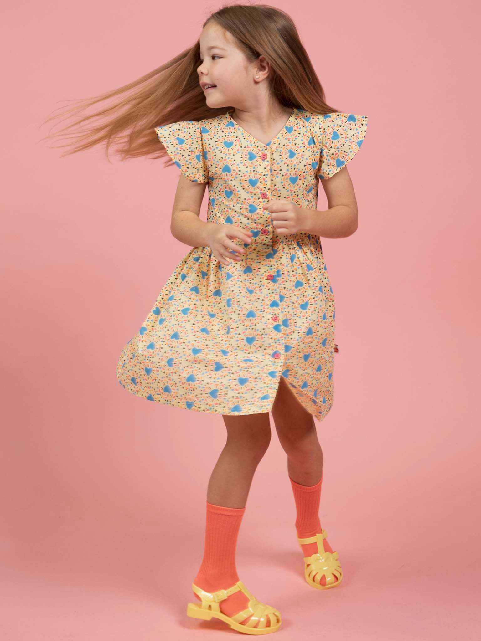 A young girl with long hair twirls in the Starburst Astra Dress by Starburst, paired with orange knee-high socks and yellow sandals, set against a pink background.