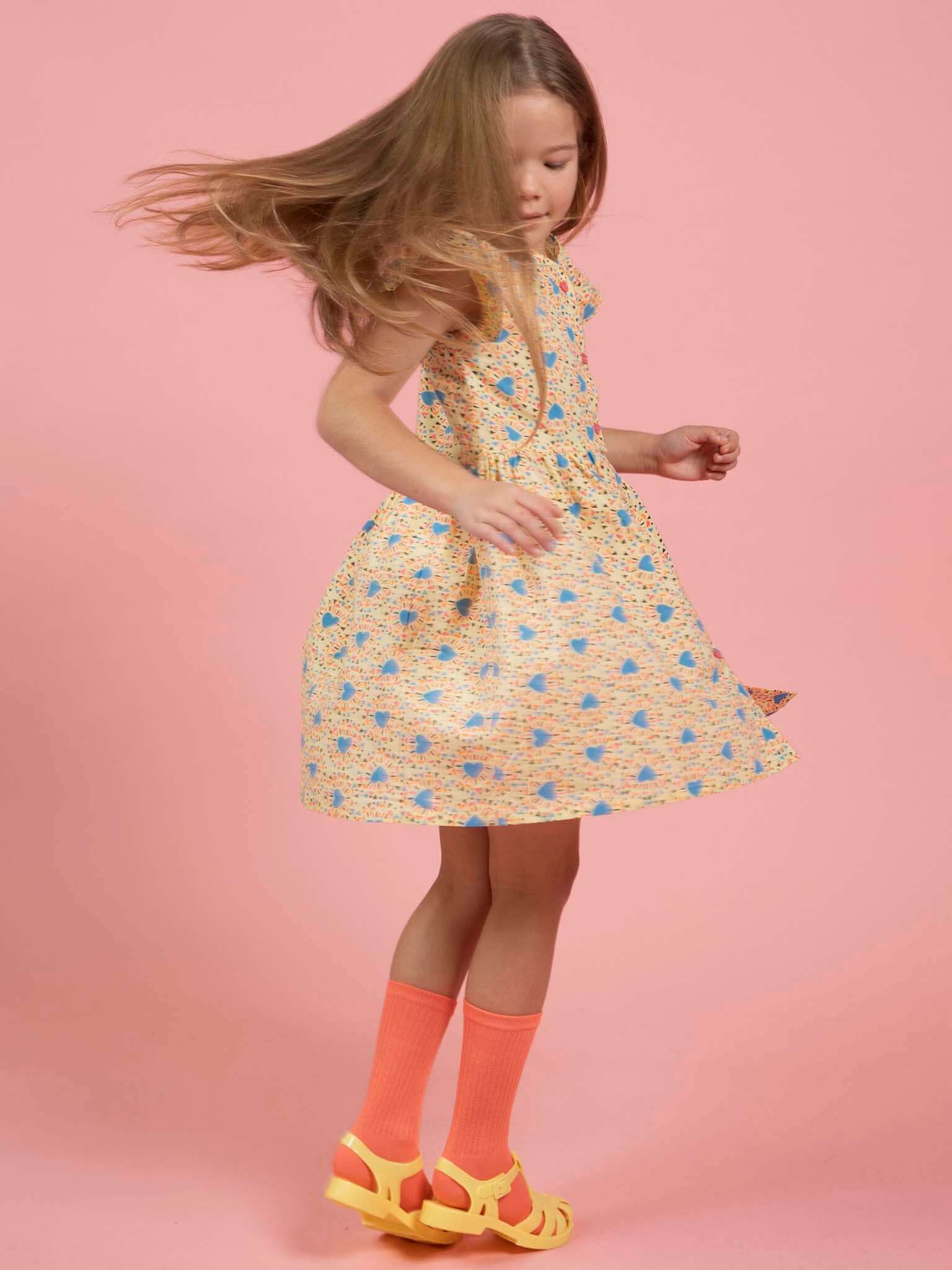 A young girl twirls in the Starburst Astra Dress by Starburst—an ethically made, sleeveless patterned dress—paired with neon orange knee-high socks and yellow sandals, all set against a plain pink background.
