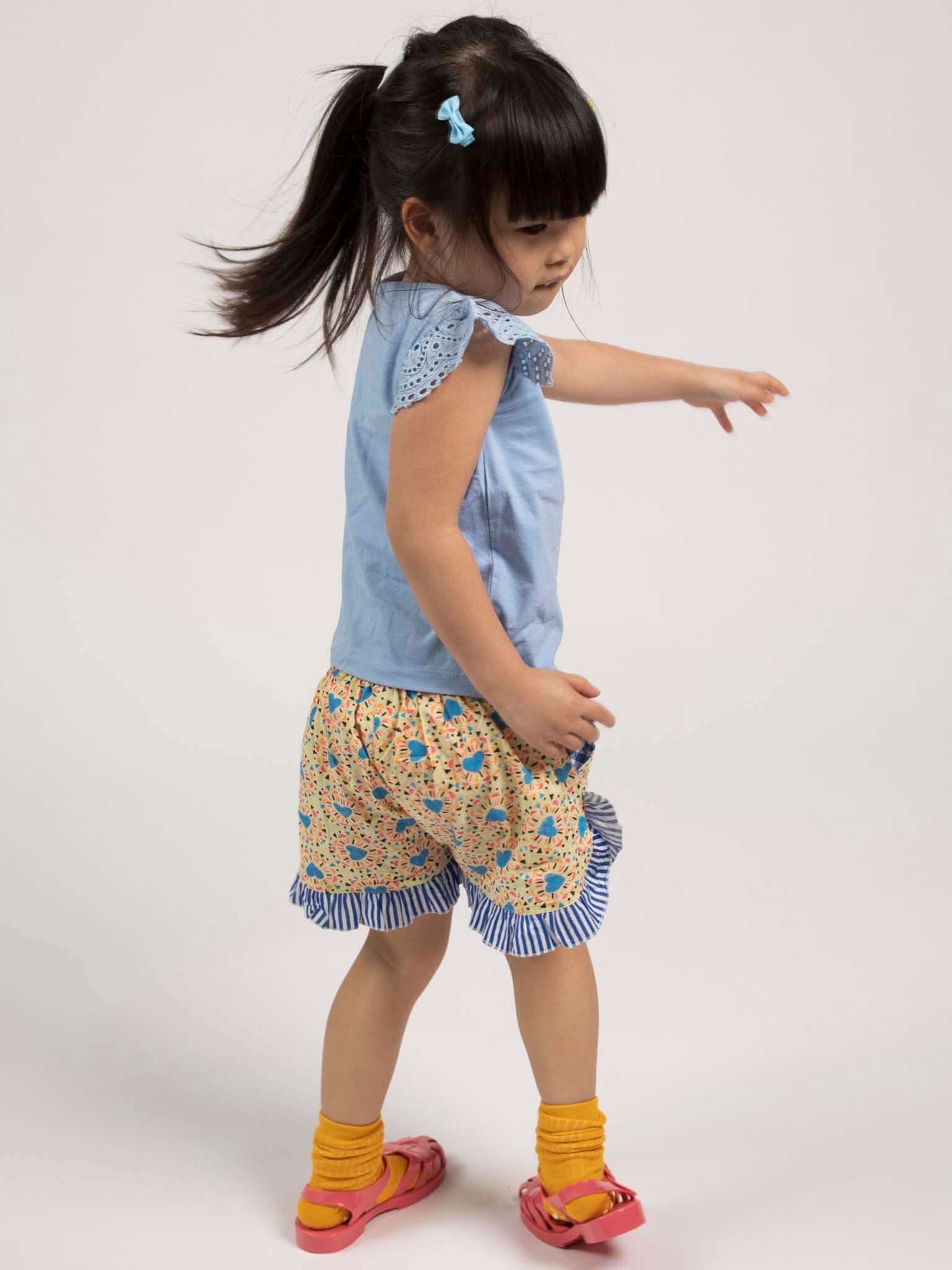 A young girl with a ponytail models Starburst Ruffle Shorts by Starburst, paired with a light blue top, yellow socks, and pink sandals. She stands and turns to her left against a plain background.