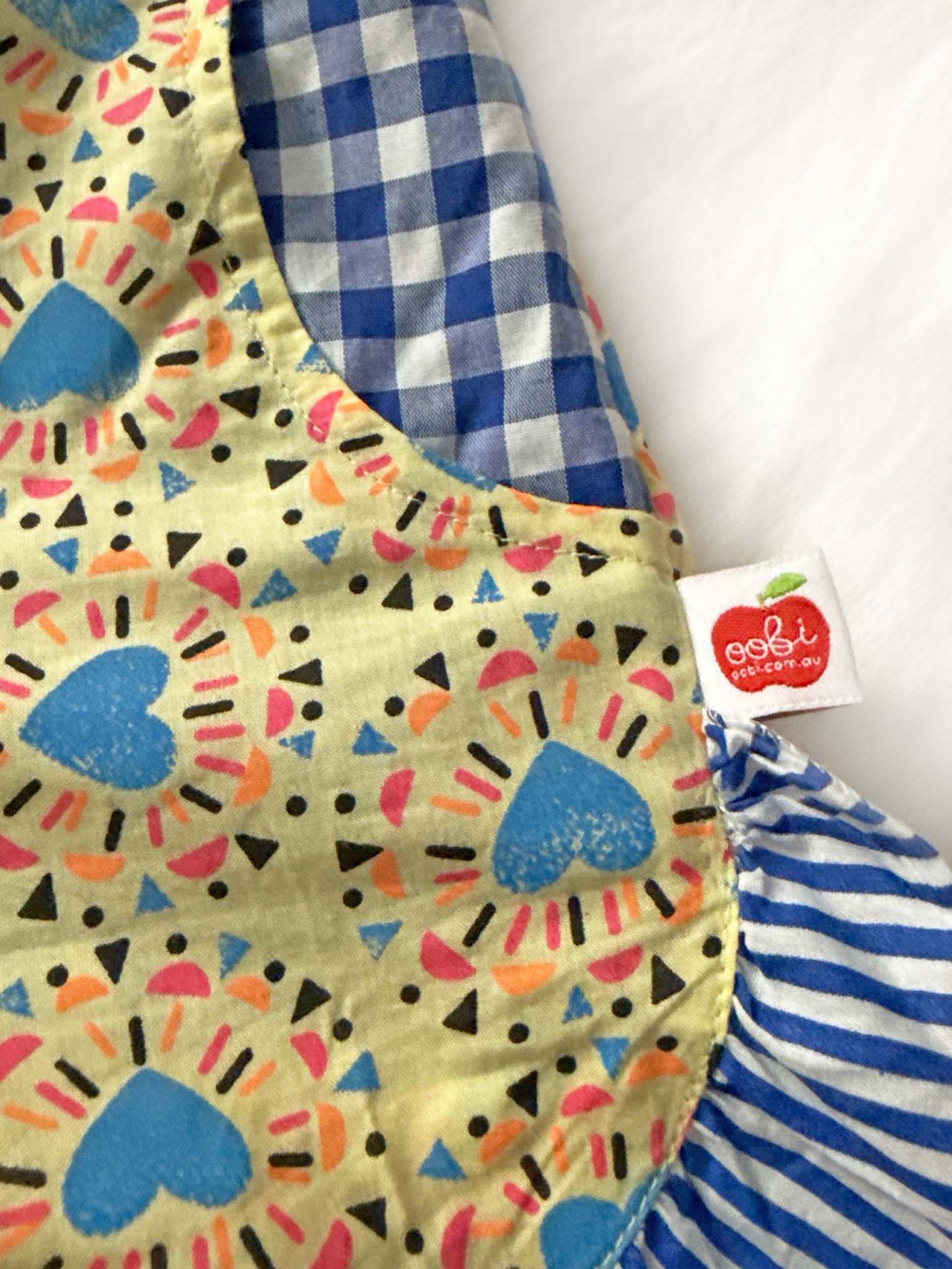 Close-up of hand screen printed fabric with blue hearts, yellow geometric shapes, checks, and stripes on Starburst Ruffle Shorts for girls, featuring a "Starburst" brand tag on the side.