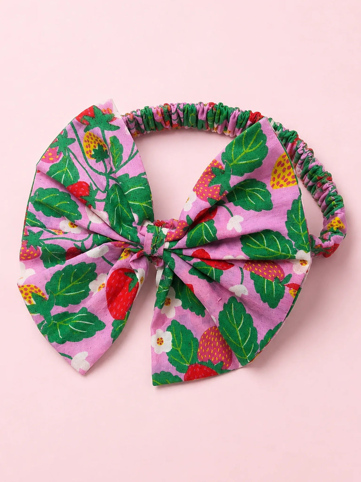 A fabric headband with a large bow, featuring a pink background with a green strawberry and leaf pattern, laid flat on a solid pink background.
