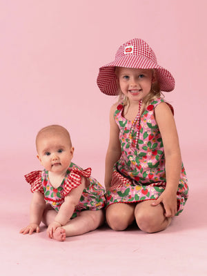 Smiling girl in the Strawberry Fields Gracie dress and gingham Chloe sun hat with her baby sister