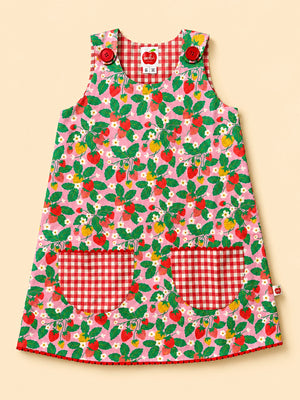 A sleeveless children's dress with a pink strawberry and leaf print, featuring red and white gingham pockets and shoulder straps, displayed on a solid beige background.