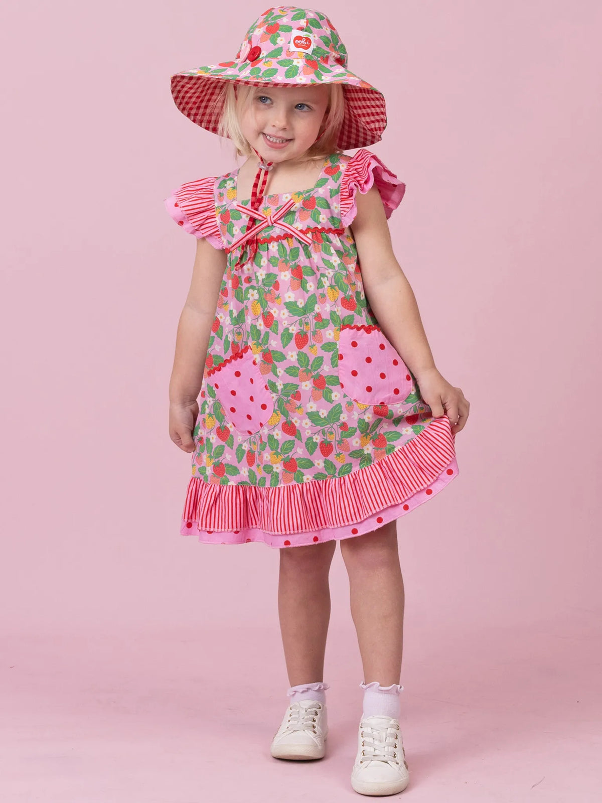 A young girl wearing a strawberry-themed dress and matching sun hat, standing against a solid pink studio background.