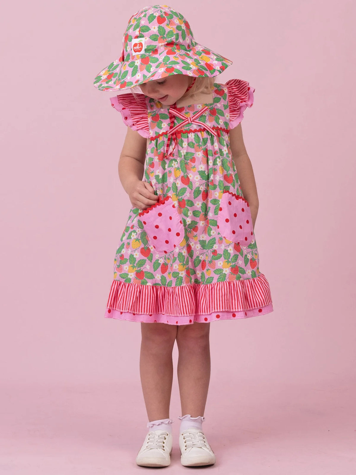 A toddler girl wearing a strawberry-themed dress and matching sun hat, standing against a solid pink studio background. The dress features a pink and white strawberry print, ruffled sleeves, a bow at the neckline, and pink strawberry-shaped pockets.