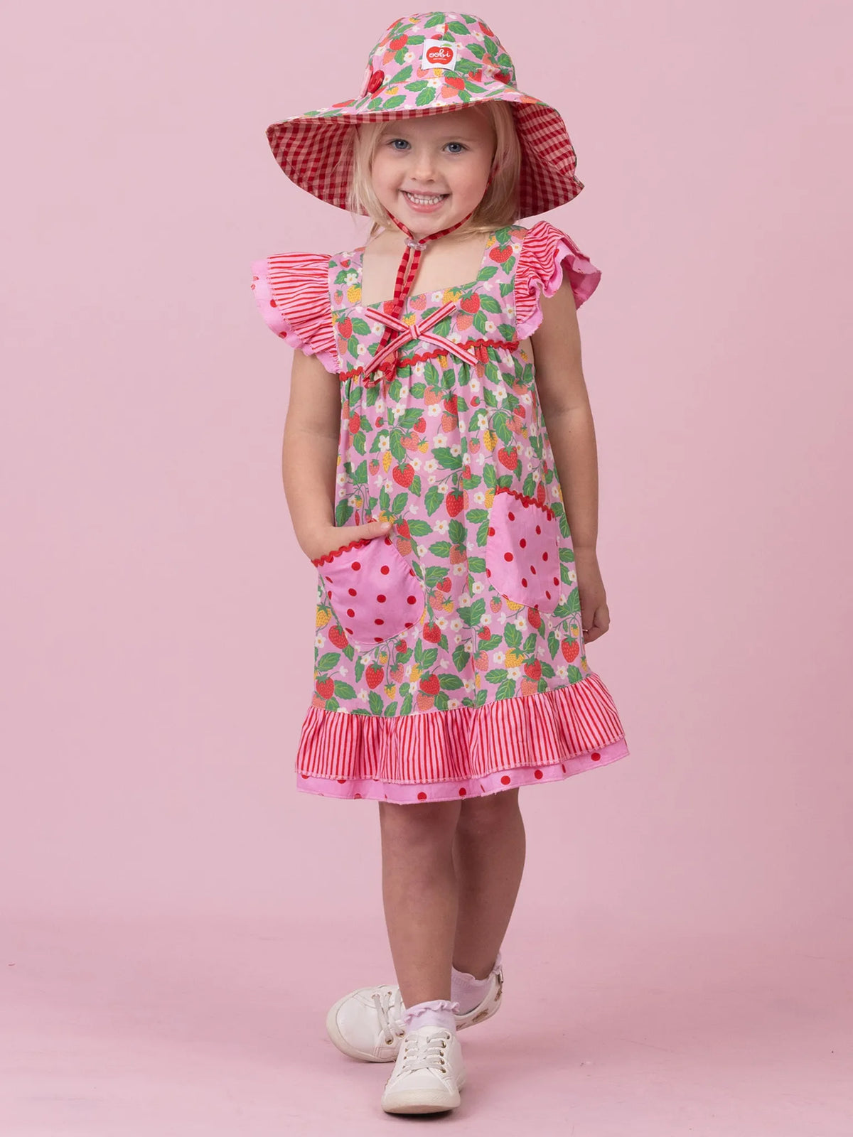A young girl wearing a strawberry-themed dress and matching sun hat, standing against a solid pink studio background.