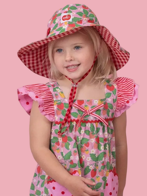 A young girl wearing a pink strawberry-print dress and a matching pink sun hat with red gingham trim, posed against a solid pink background.