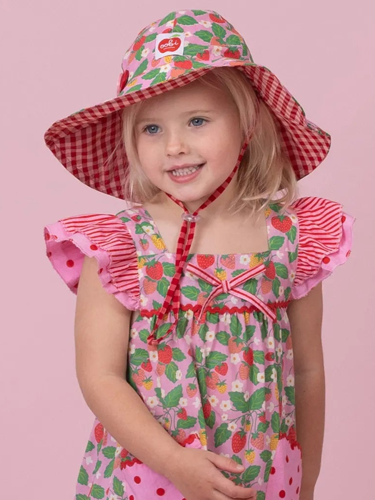 A young girl wearing a pink strawberry-print dress and a matching pink sun hat with red gingham trim, posed against a solid pink background.