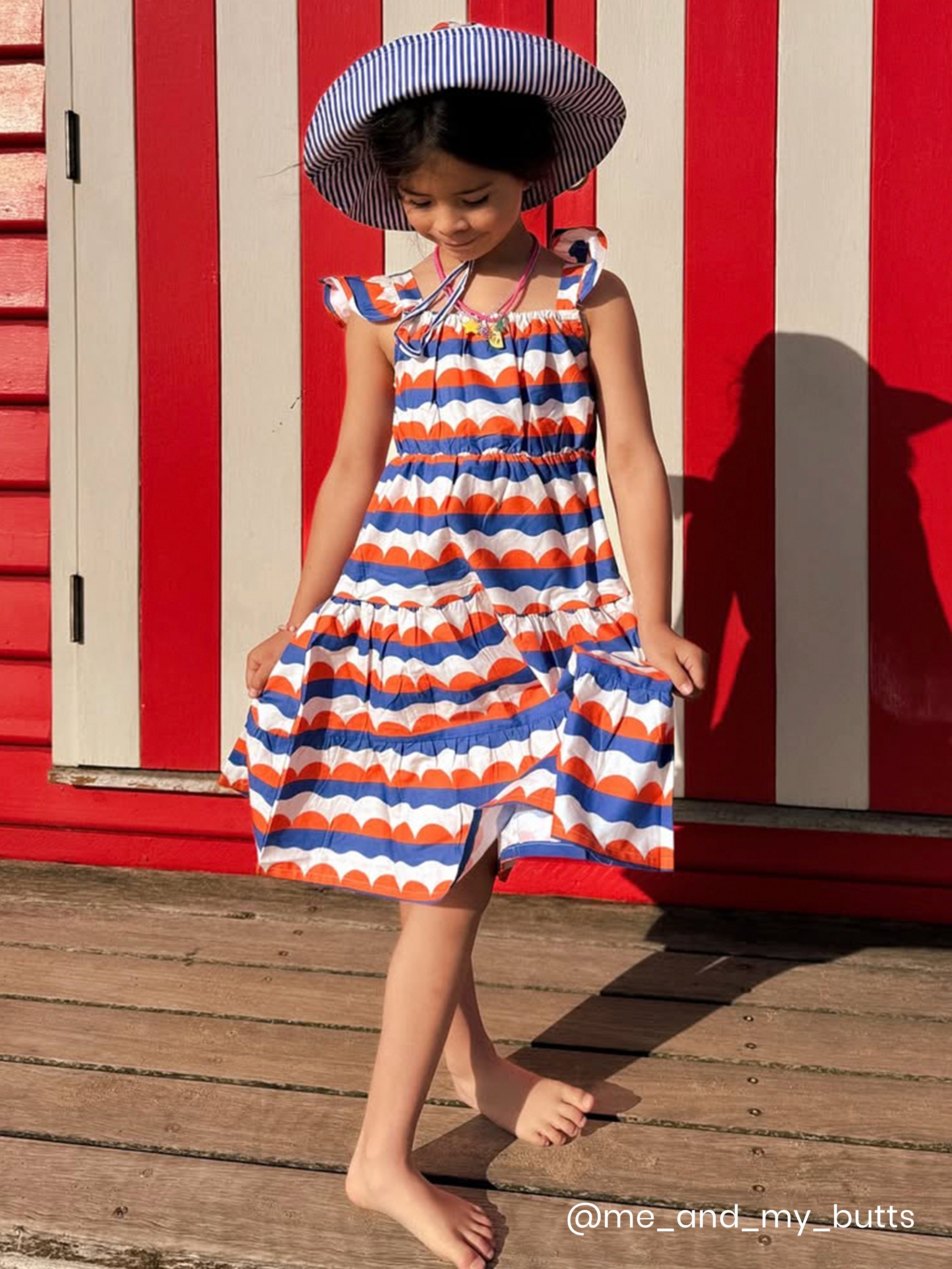 Young girl in a striped dress and sun hat standing on a wooden deck with a red and white striped wall in the background.