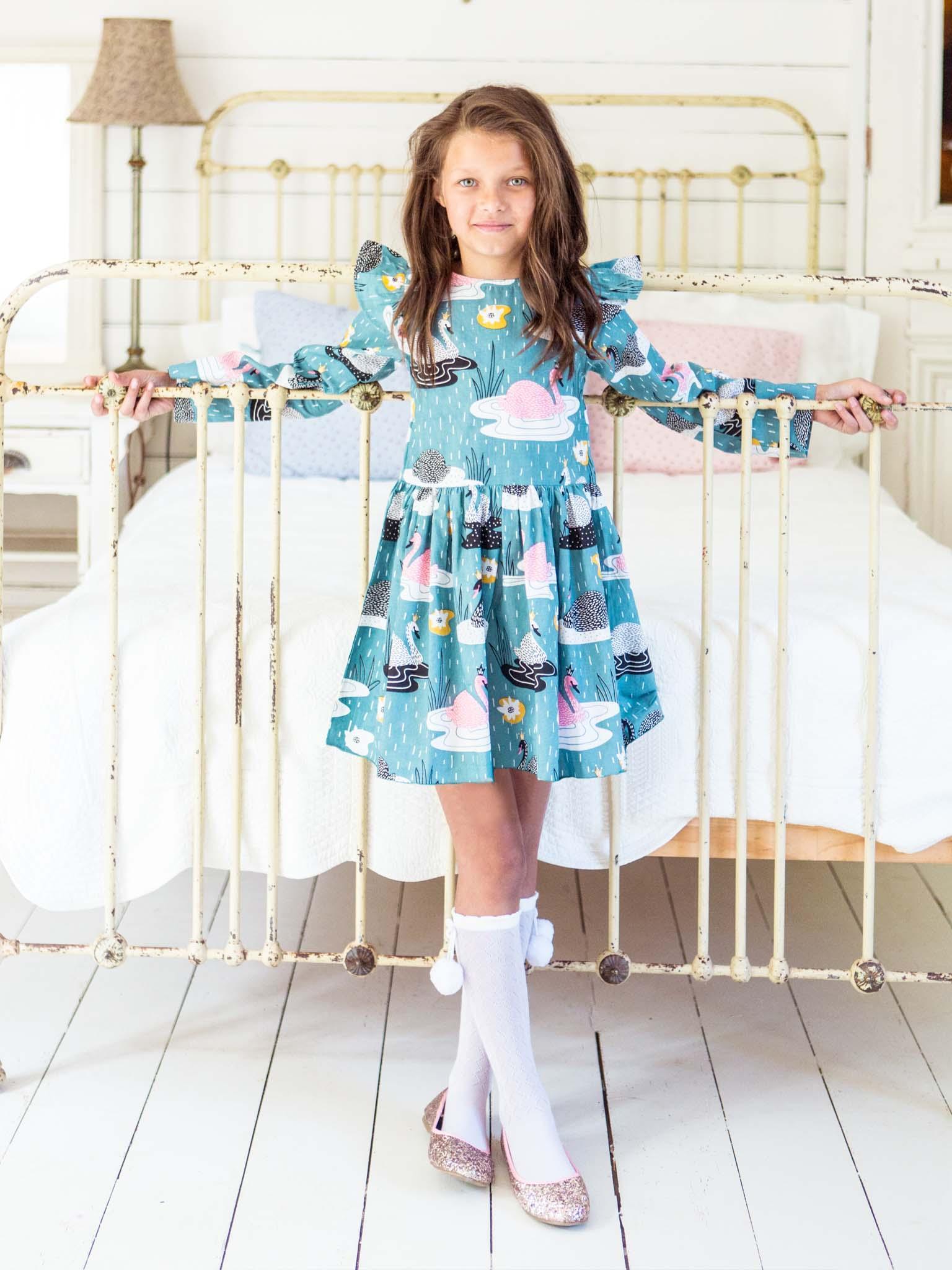 Young girl in a Swan Song Dahlia Dress and sparkly shoes standing by an antique metal bed in a brightly lit room.