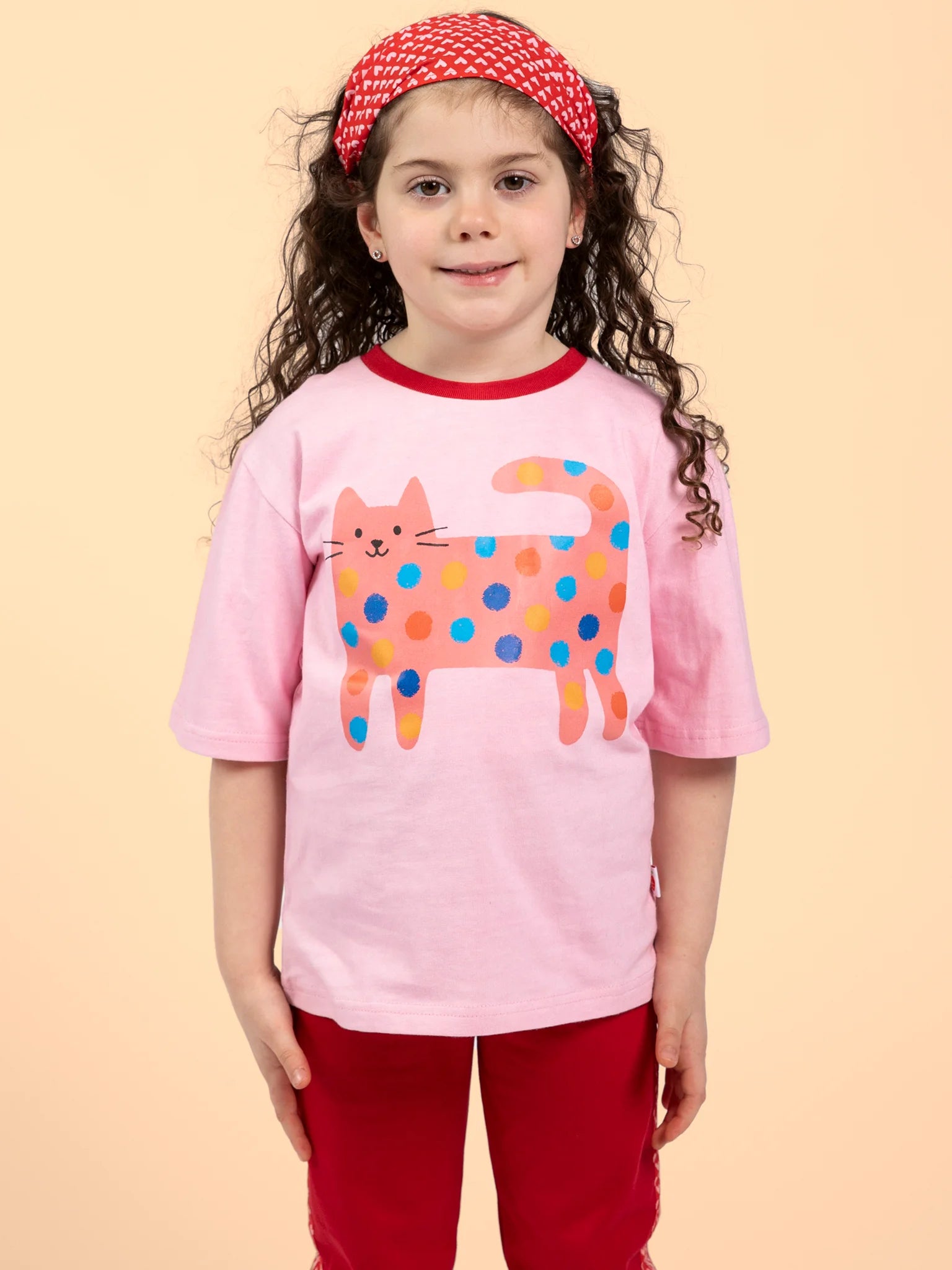 Young girl with long curly hair and a red headband wears the Essentials Quirky Cat Pink Tee with Red Binding and red pants, standing against a plain light background.