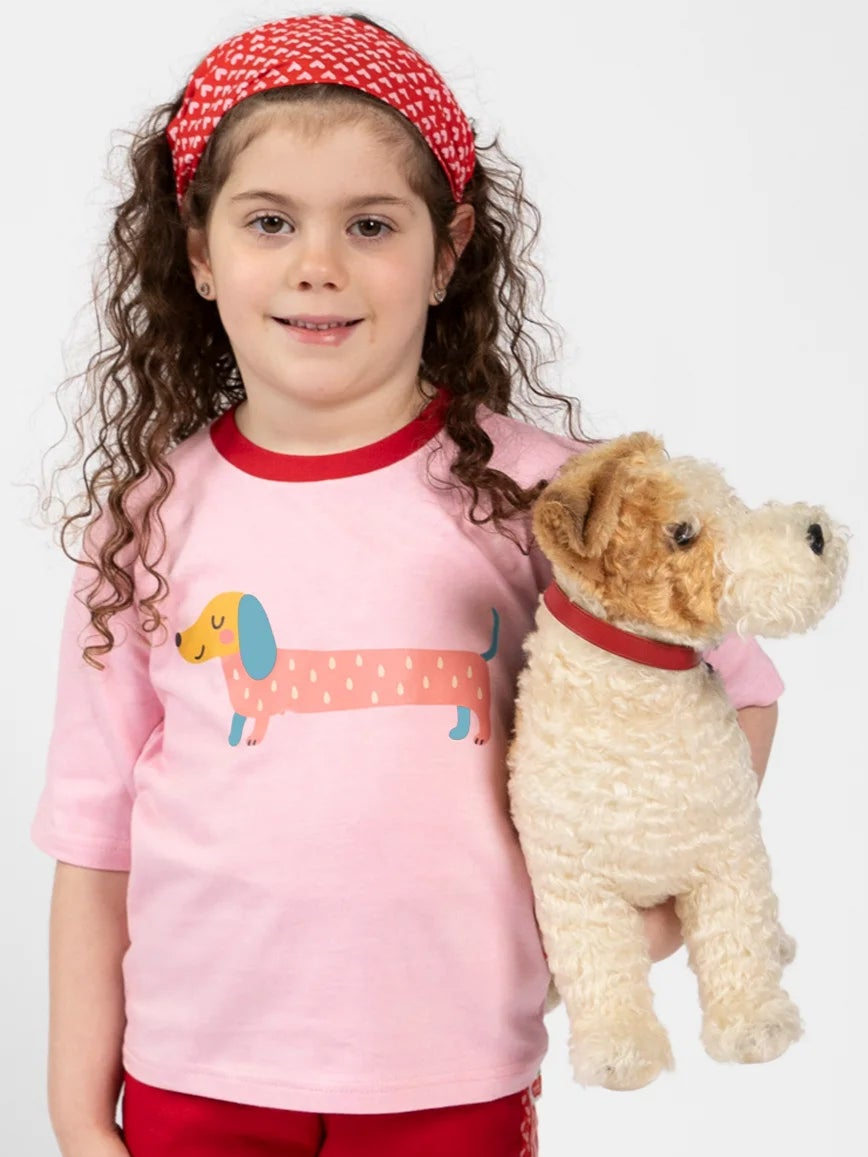 A young girl wears the Essentials Pink Sausage Dog Tee with Red Binding, styled oversized. She accessorizes with a red headband and holds a plush dog toy sporting a red collar.