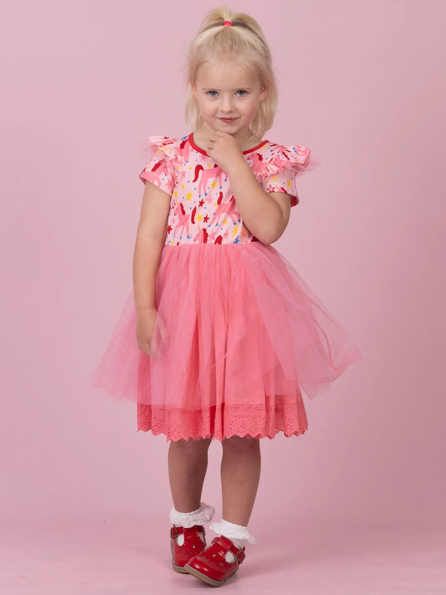 A young blonde girl wearing a pink and white patterned dress with a pink tulle skirt, standing against a solid pink studio background.