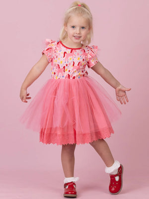 A young girl with blonde hair in a ponytail is wearing a pink tulle tutu dress with a colorful floral pattern bodice, posing against a solid pink background.