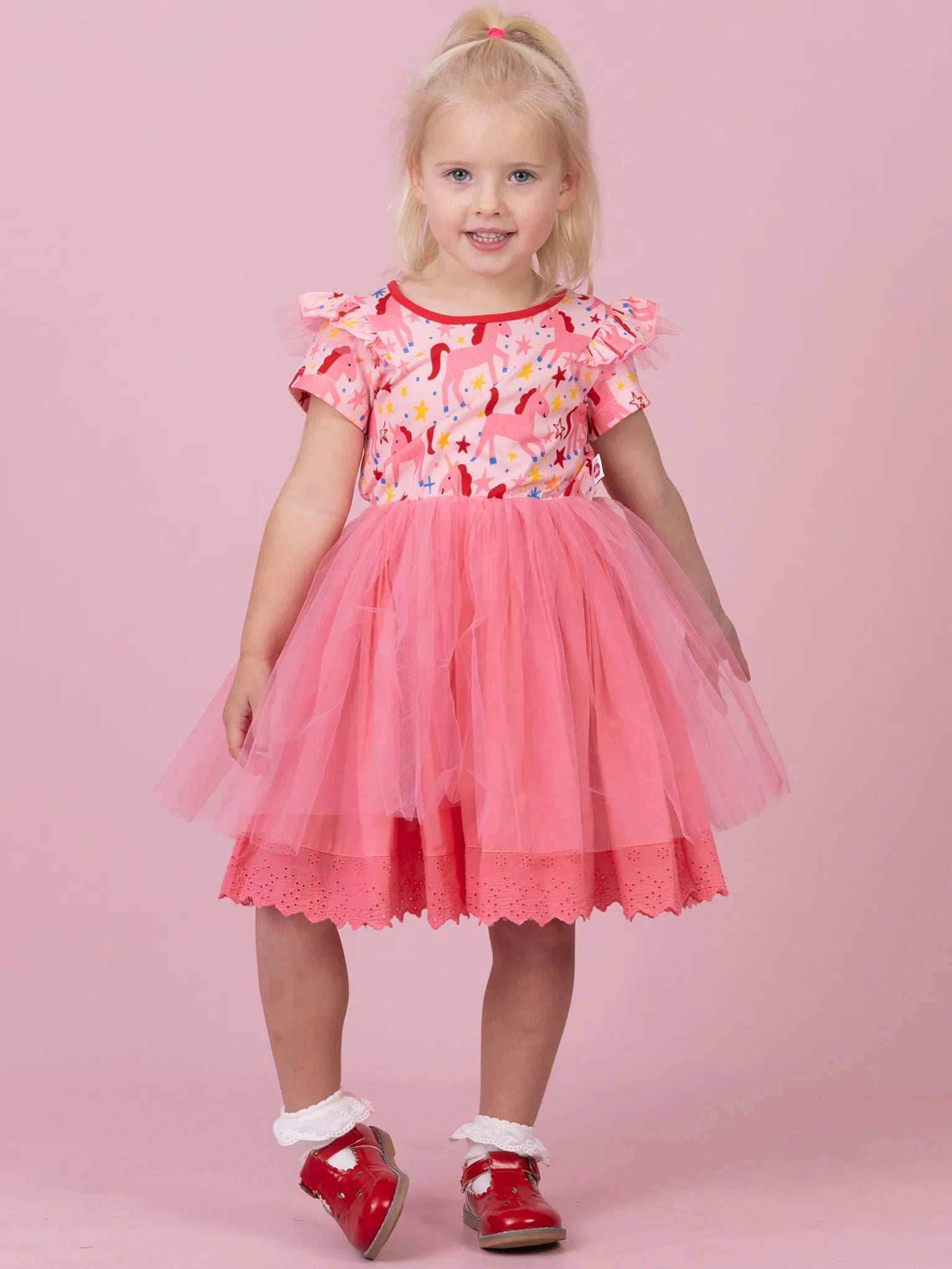 A young girl with blonde hair in a ponytail stands against a solid pink studio background, wearing a pink and red dress with a floral pattern bodice, ruffled sleeves, and a layered tulle skirt, paired with red shoes and white socks.