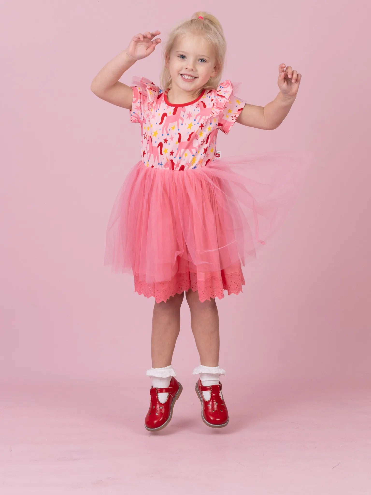 A young girl wearing a pink and red candy-themed tutu dress with white socks and red shoes, posing against a solid pink studio background.