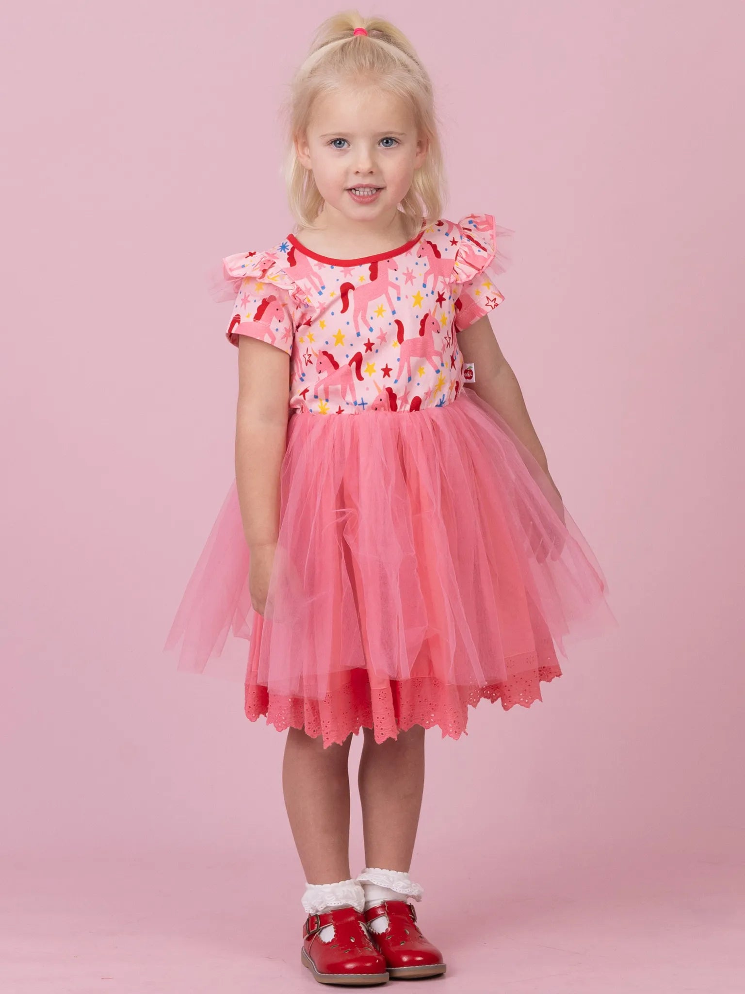 A young girl with blonde hair in a ponytail stands against a solid pink studio background, wearing a pink dress with a floral pattern bodice, ruffled short sleeves, and a layered tulle skirt, paired with red shoes.