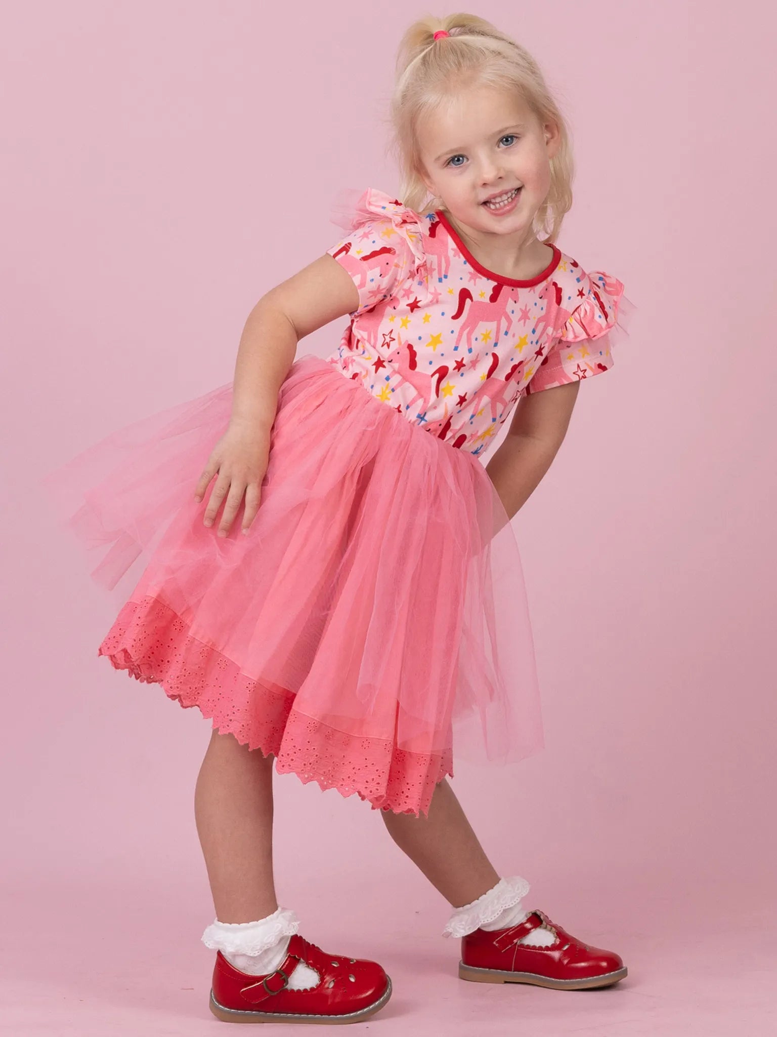 A young girl modeling a pink tulle dress with a floral bodice, standing against a solid pink studio background. She is wearing red Mary Jane shoes and white socks.