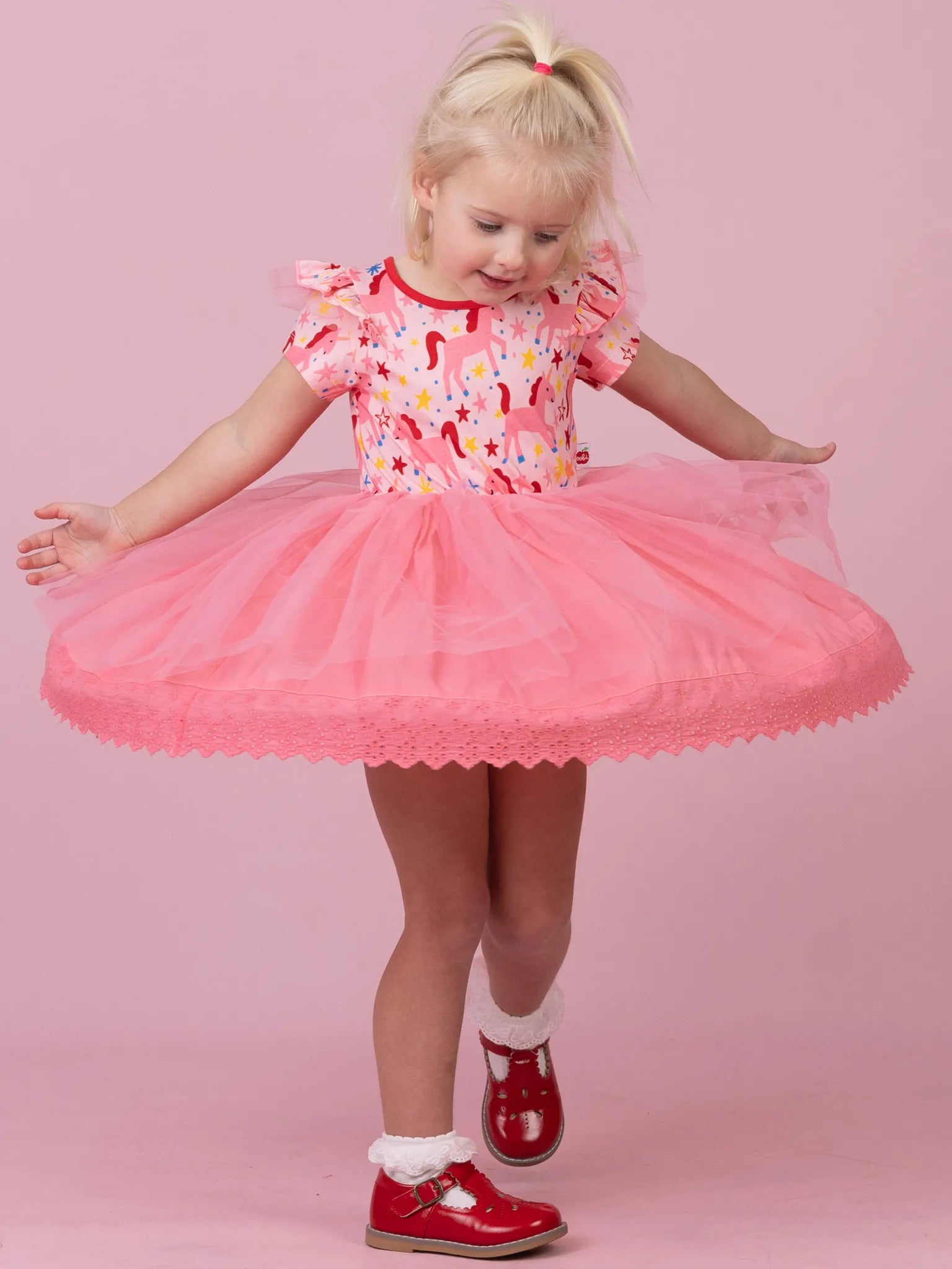 A young blonde girl wearing a pink tutu dress with a colorful floral bodice, posing against a solid pink studio background.