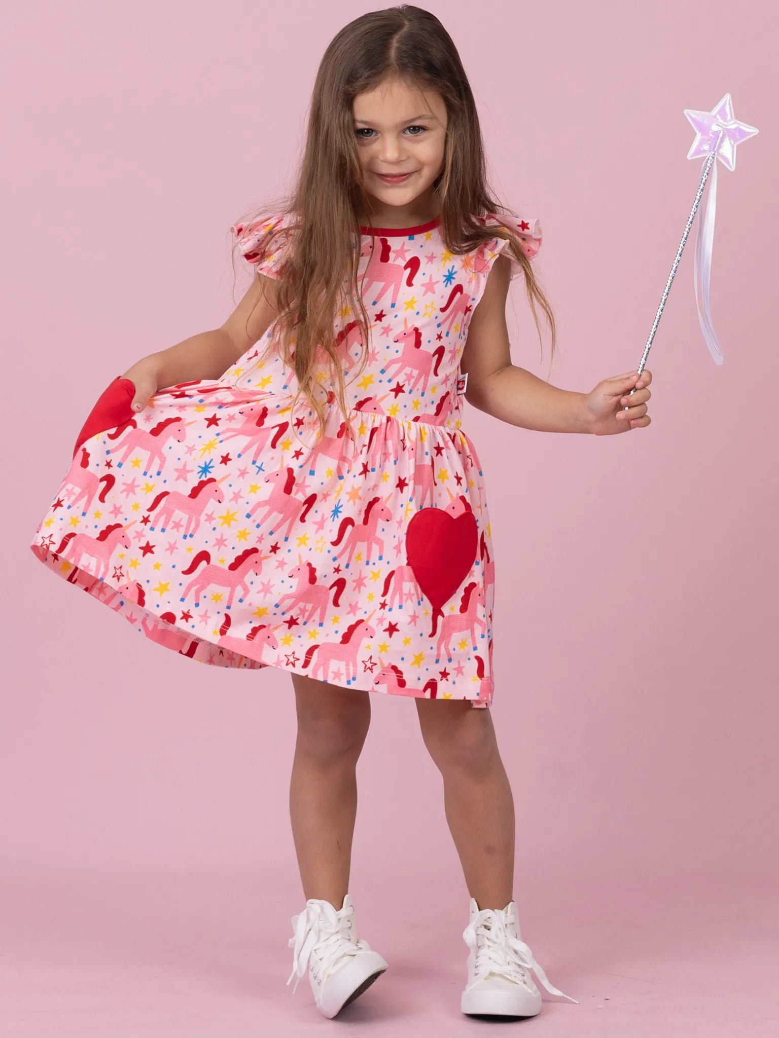 A young girl with long brown hair stands against a solid pink studio background, wearing a pink dress with a red, yellow, and blue unicorn print, red heart-shaped pockets, and holding a silver star wand.