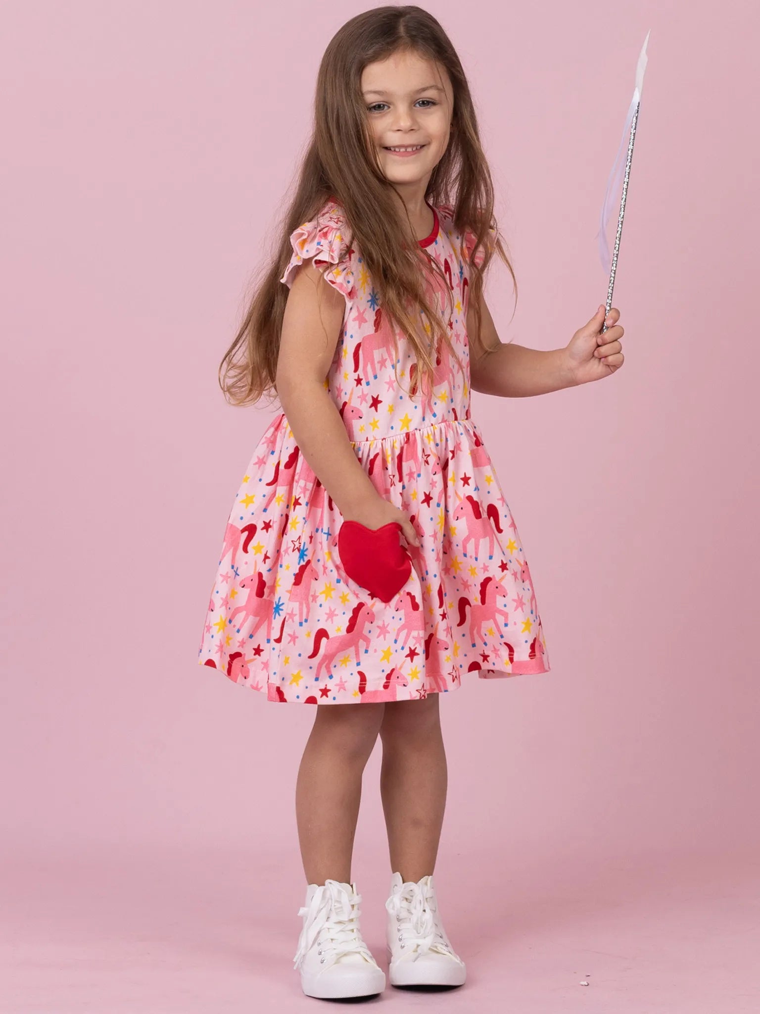 A young girl with long brown hair stands against a solid pink studio background, wearing a pink dress with a multicolor unicorn and floral print, holding a silver wand, and wearing white sneakers.