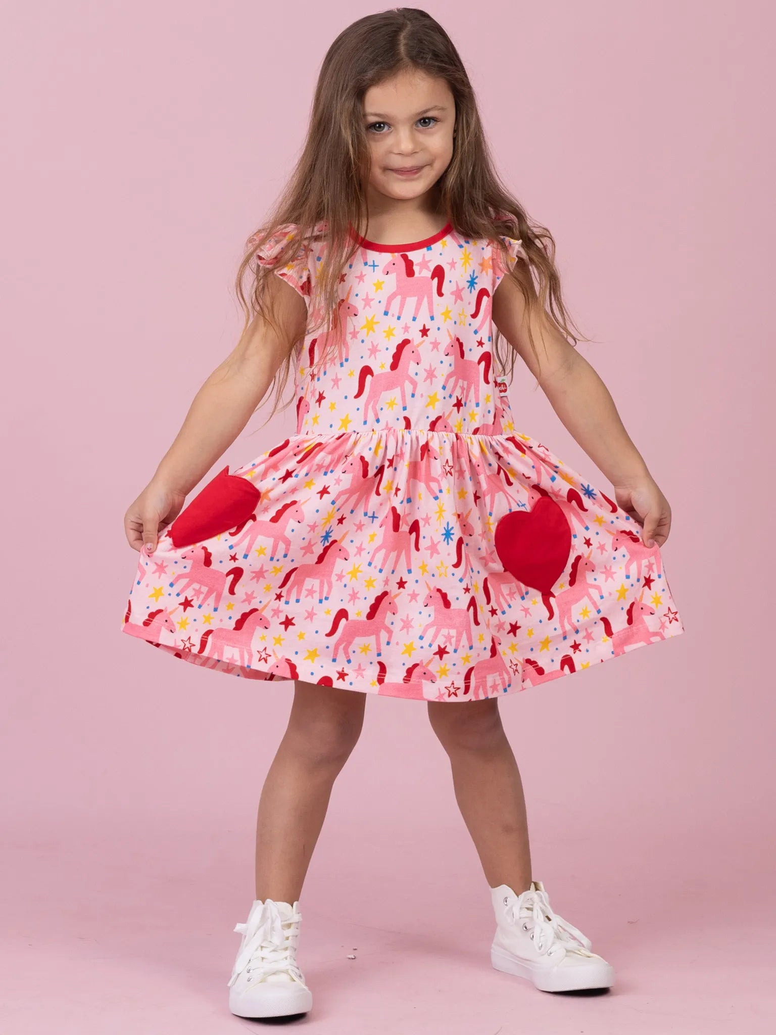A young girl with long brown hair stands against a solid pink studio background wearing a pink unicorn-print dress with red heart-shaped pockets and white sneakers.