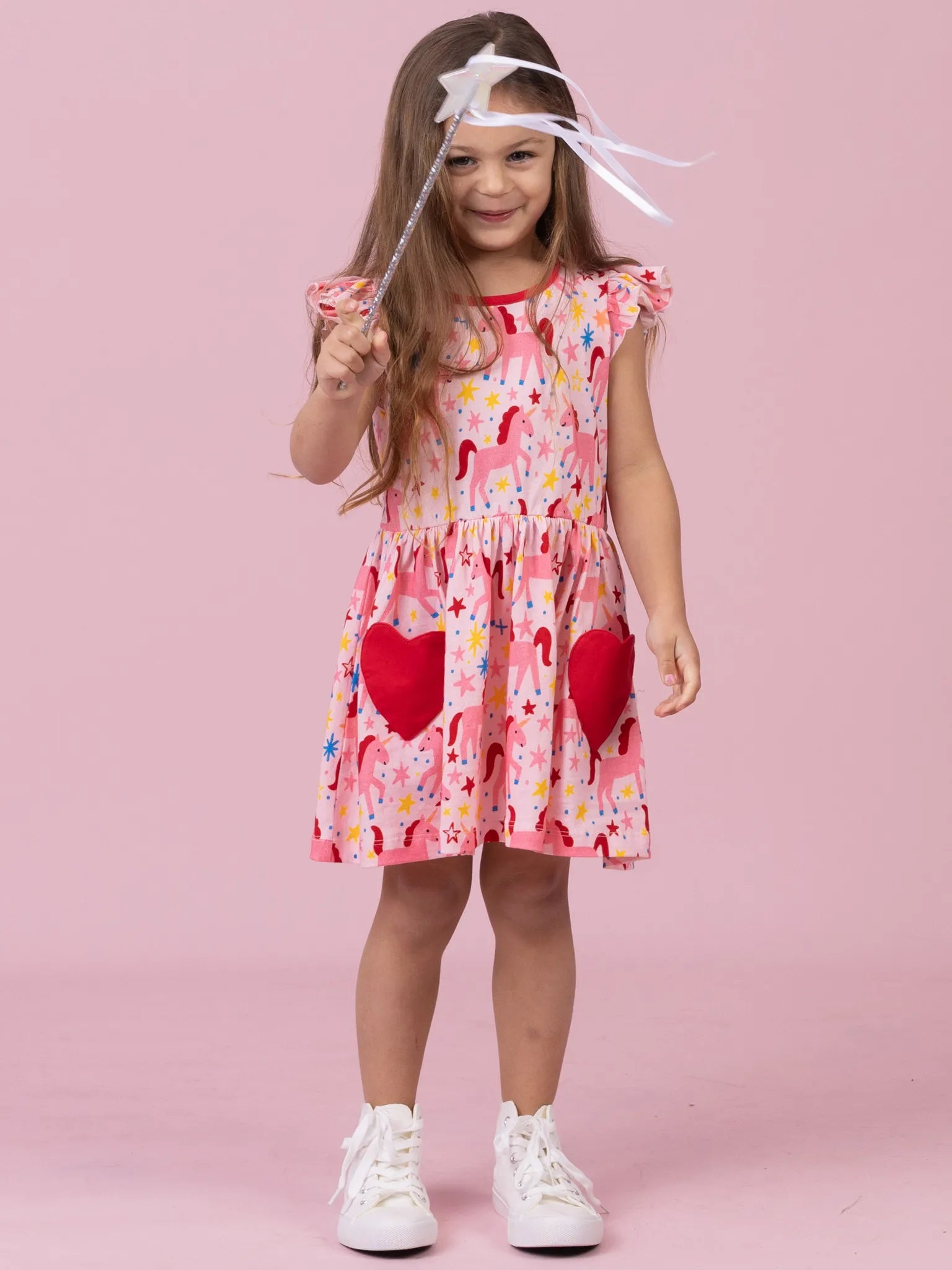 A young girl with long brown hair stands against a solid pink studio background, wearing a pink dress with a colorful print, red heart-shaped pockets, and white sneakers. She is holding a white wand with a star topper.