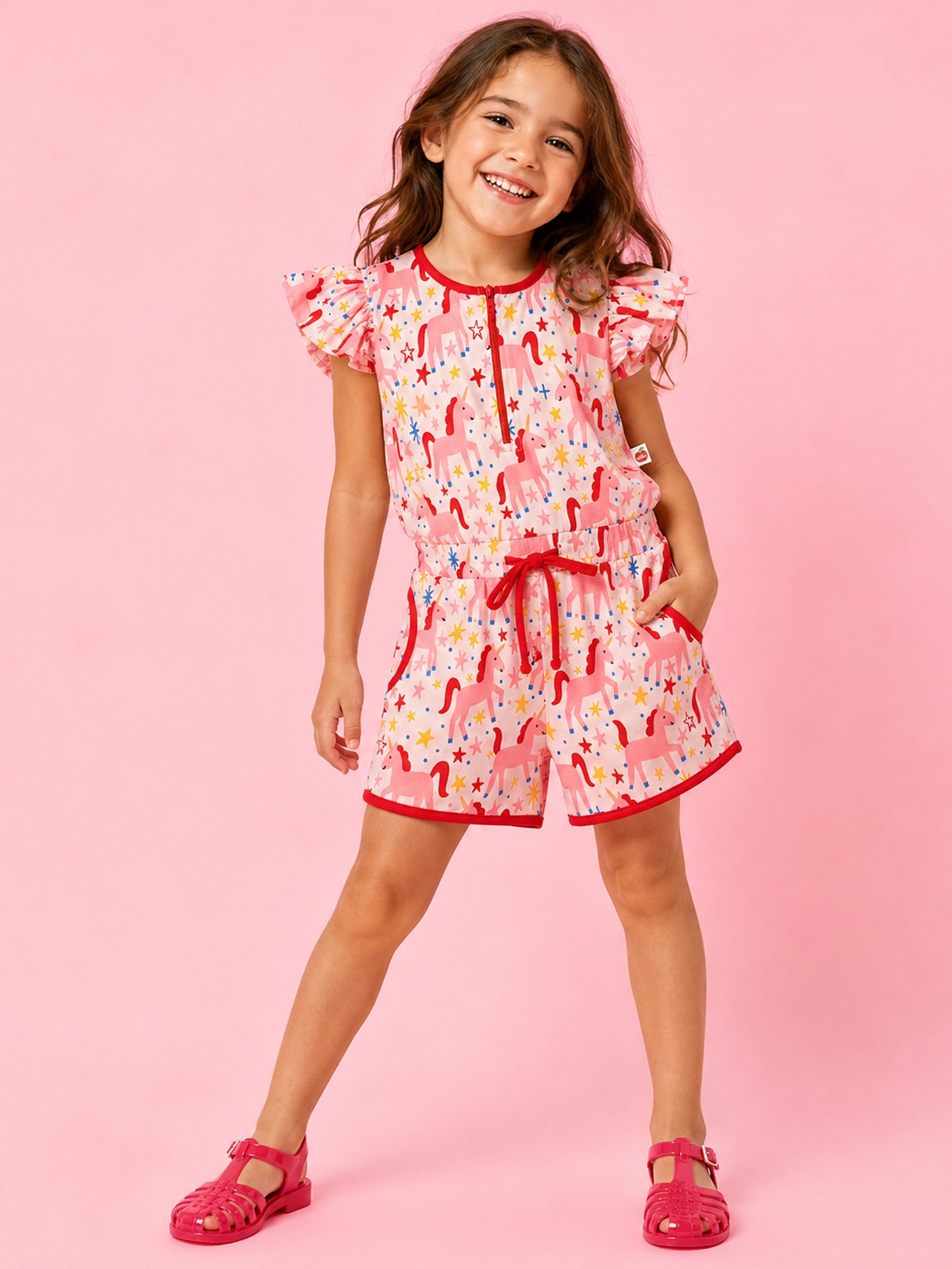 A young girl modeling a pink and white unicorn-themed romper with ruffled sleeves and a matching pink shoe, set against a solid pink studio background.