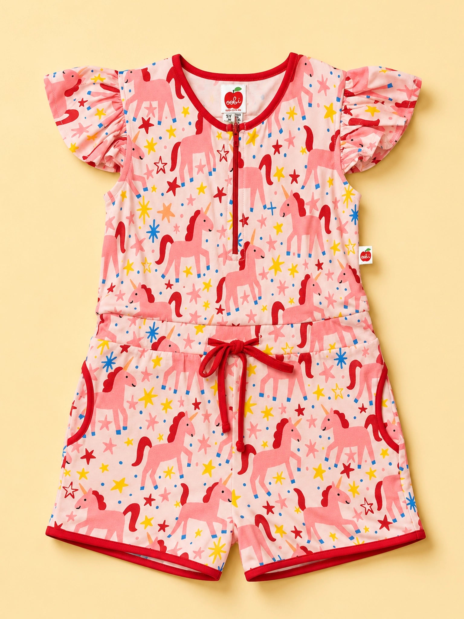A baby romper set featuring a pink unicorn and star pattern, with red trim, flutter sleeves, a front zipper, and a matching drawstring waist, laid flat on a light yellow background.