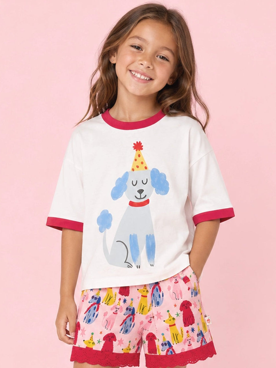 A young girl stands against a solid pink studio background wearing a white ringer T-shirt with red trim and a blue poodle graphic, paired with pink patterned shorts featuring red scalloped hems, white socks, and white low-top sneakers.