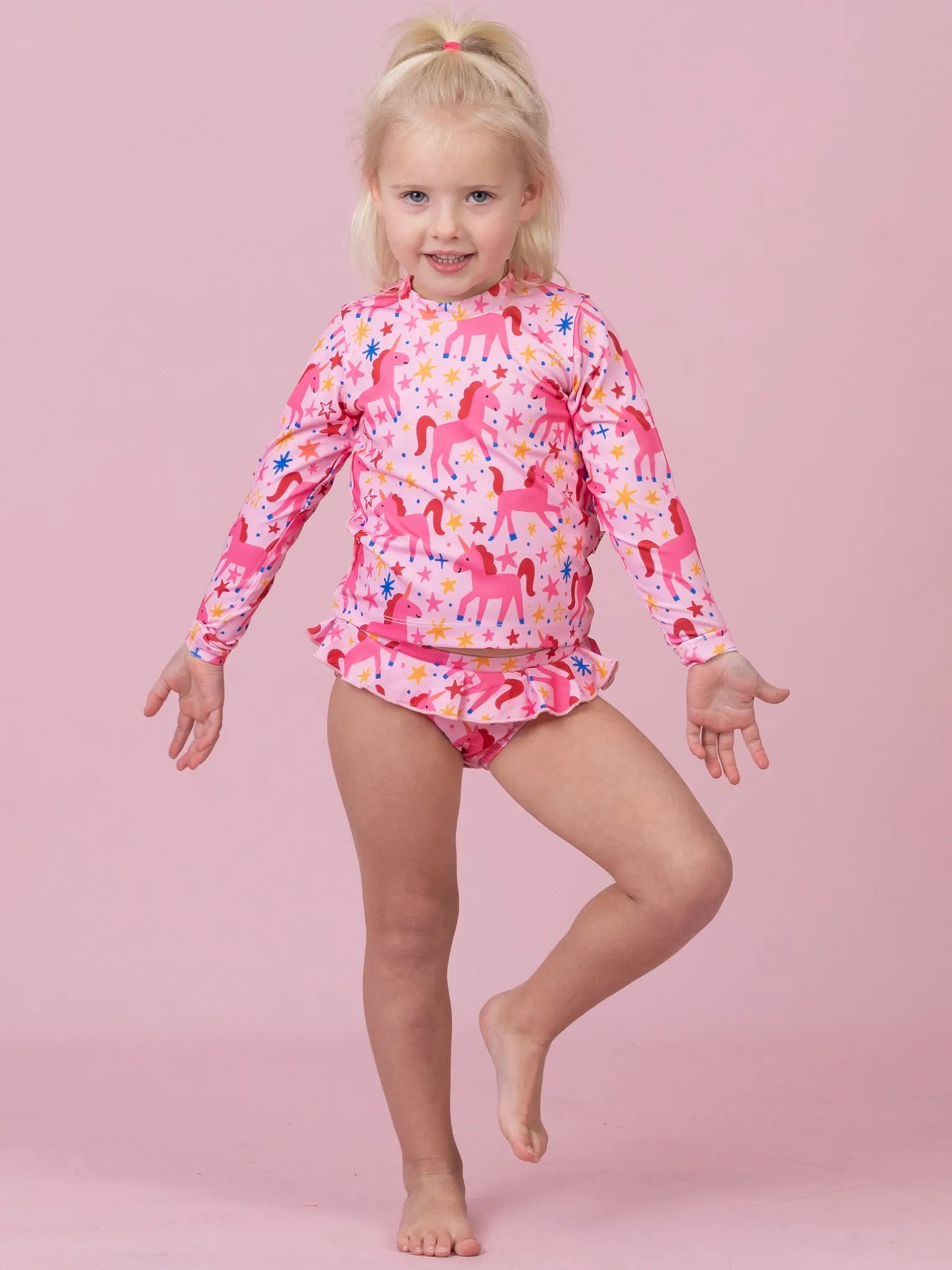 A young blonde girl wearing a pink long-sleeve swim shirt and matching swim briefs with a colorful unicorn pattern, posing against a solid pink background.