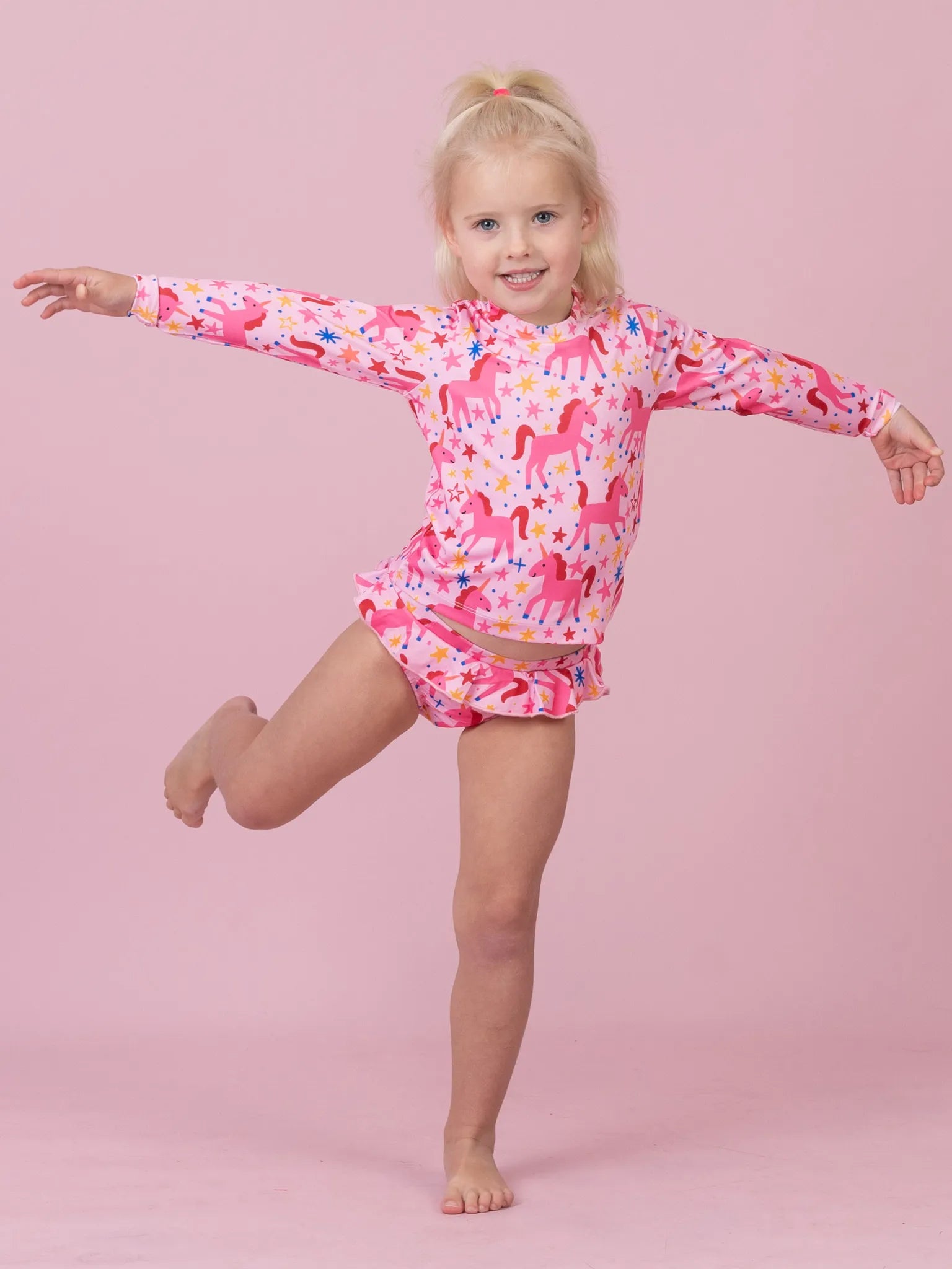 A young girl with blonde hair in a ponytail poses in a pink long-sleeve swim shirt and matching swim shorts featuring a colorful unicorn pattern, set against a solid pink studio background.
