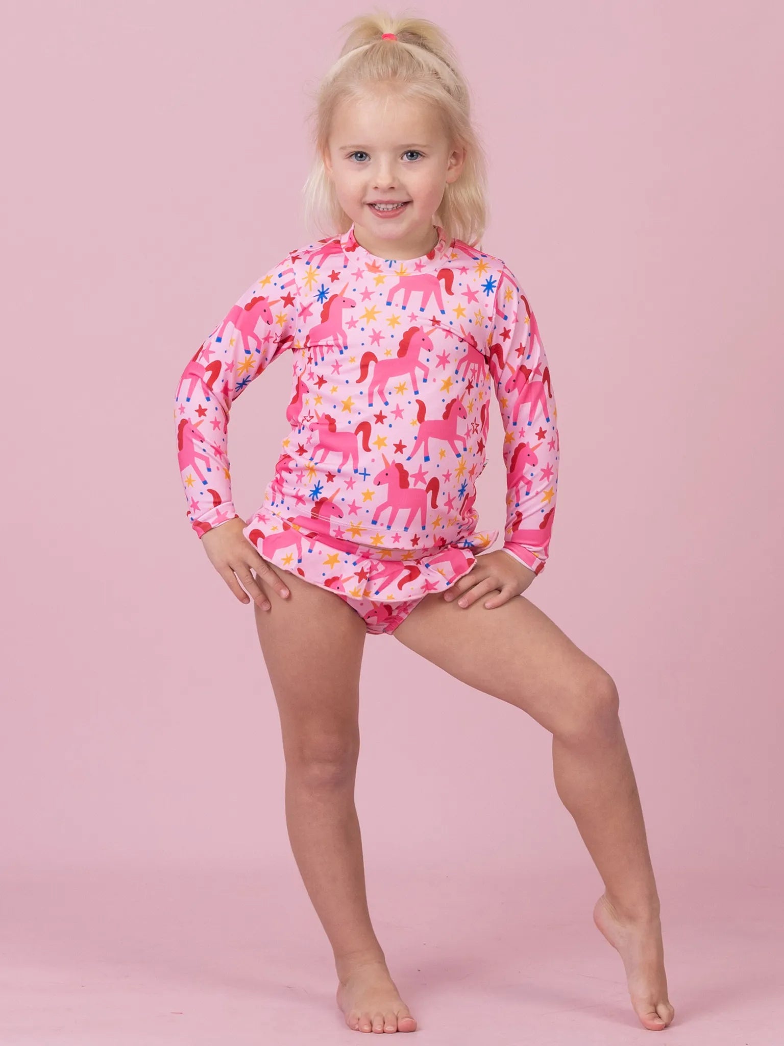 A young blonde girl modeling a pink long-sleeve swim shirt and matching swim briefs with a colorful unicorn pattern, posed against a solid pink studio background.