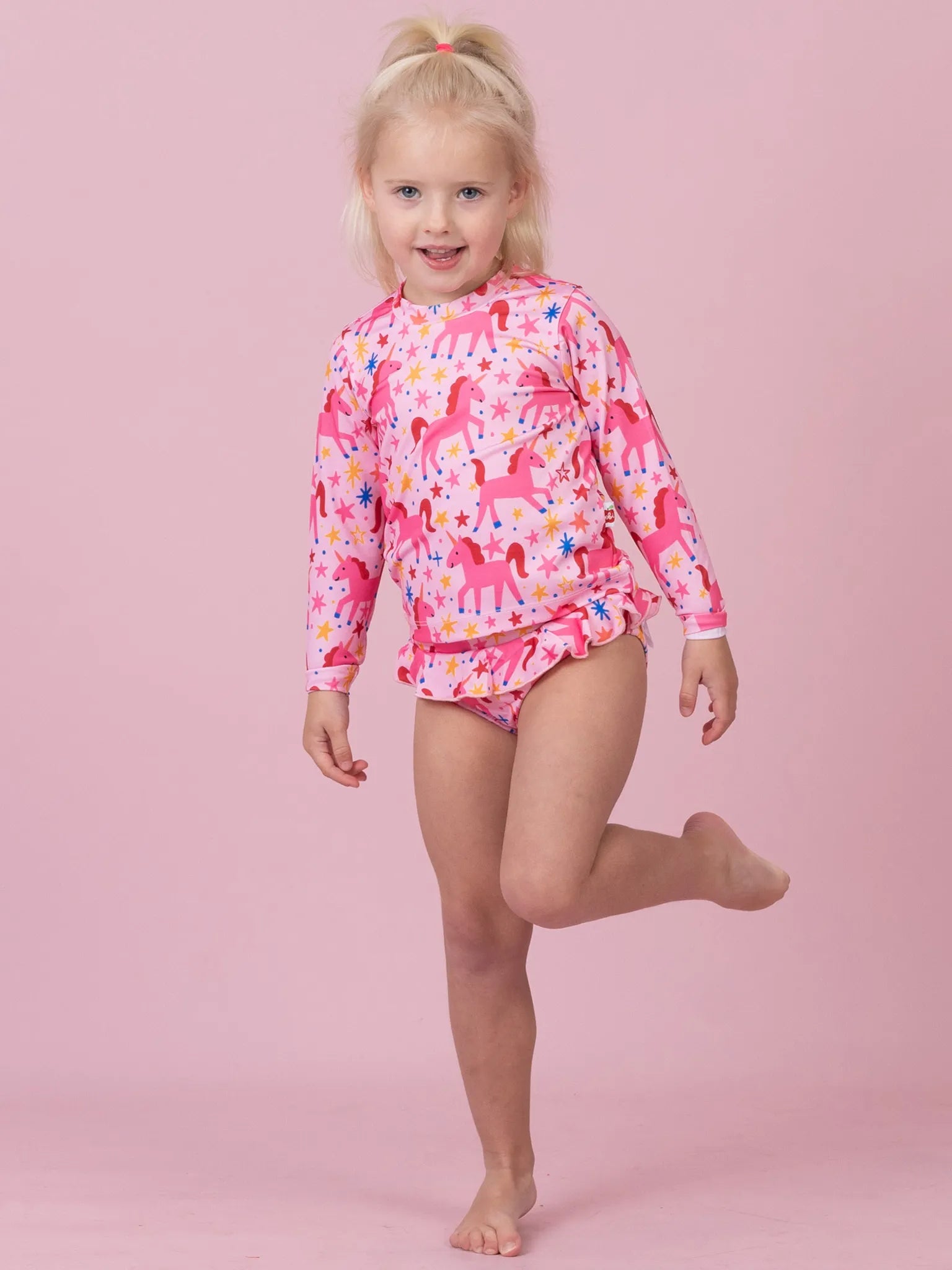 A young blonde girl wearing a pink long-sleeve swim shirt with a unicorn pattern and matching swim briefs, posing against a solid pink background.