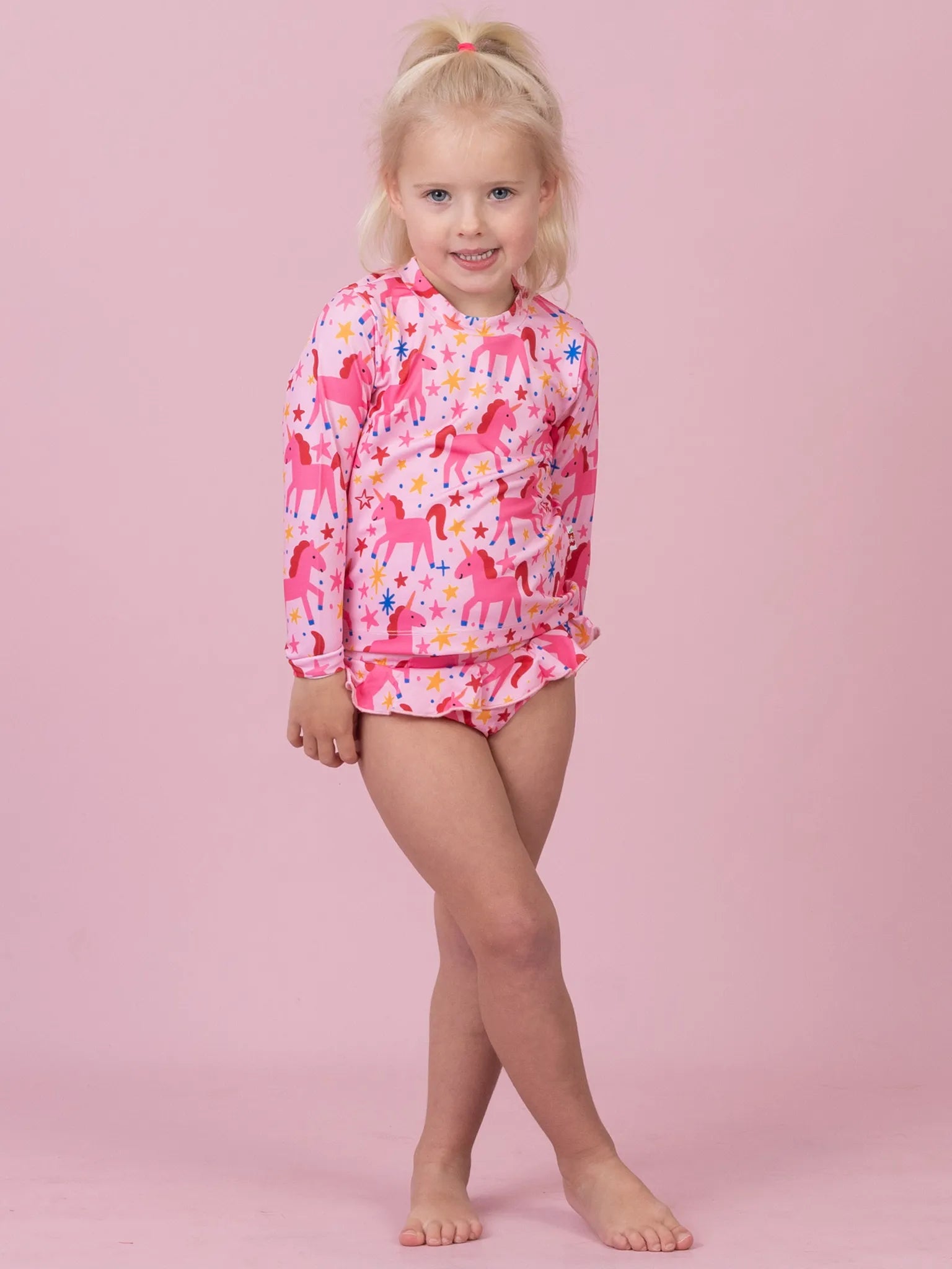 A young girl with blonde hair in a ponytail stands barefoot against a solid pink studio background, wearing a long-sleeve pink swim shirt and matching swim bottoms covered in a colorful unicorn print.