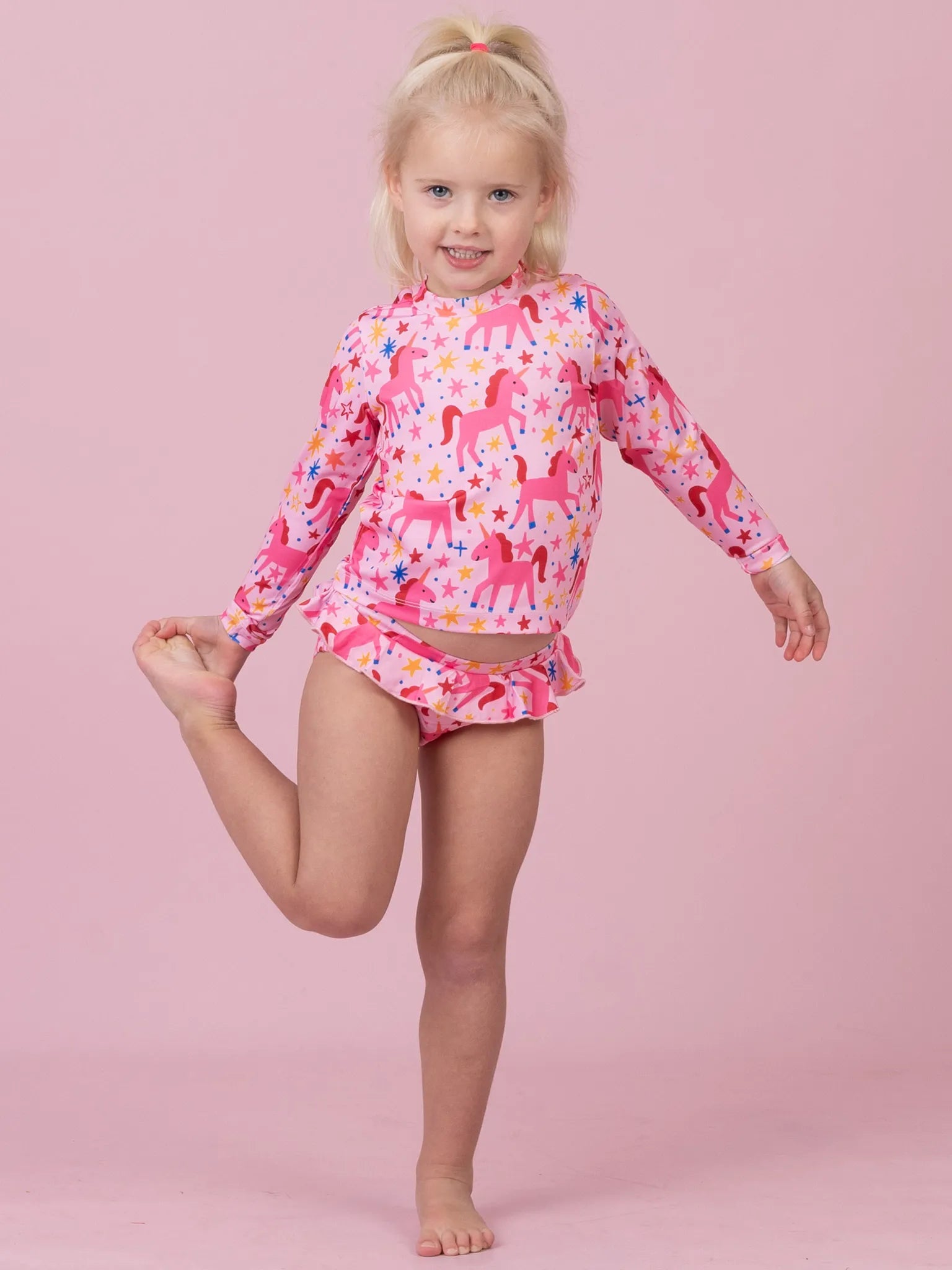 A young blonde girl posing in a pink long-sleeve swim shirt and matching swim trunks set, both featuring a colorful unicorn pattern, against a solid pink background.