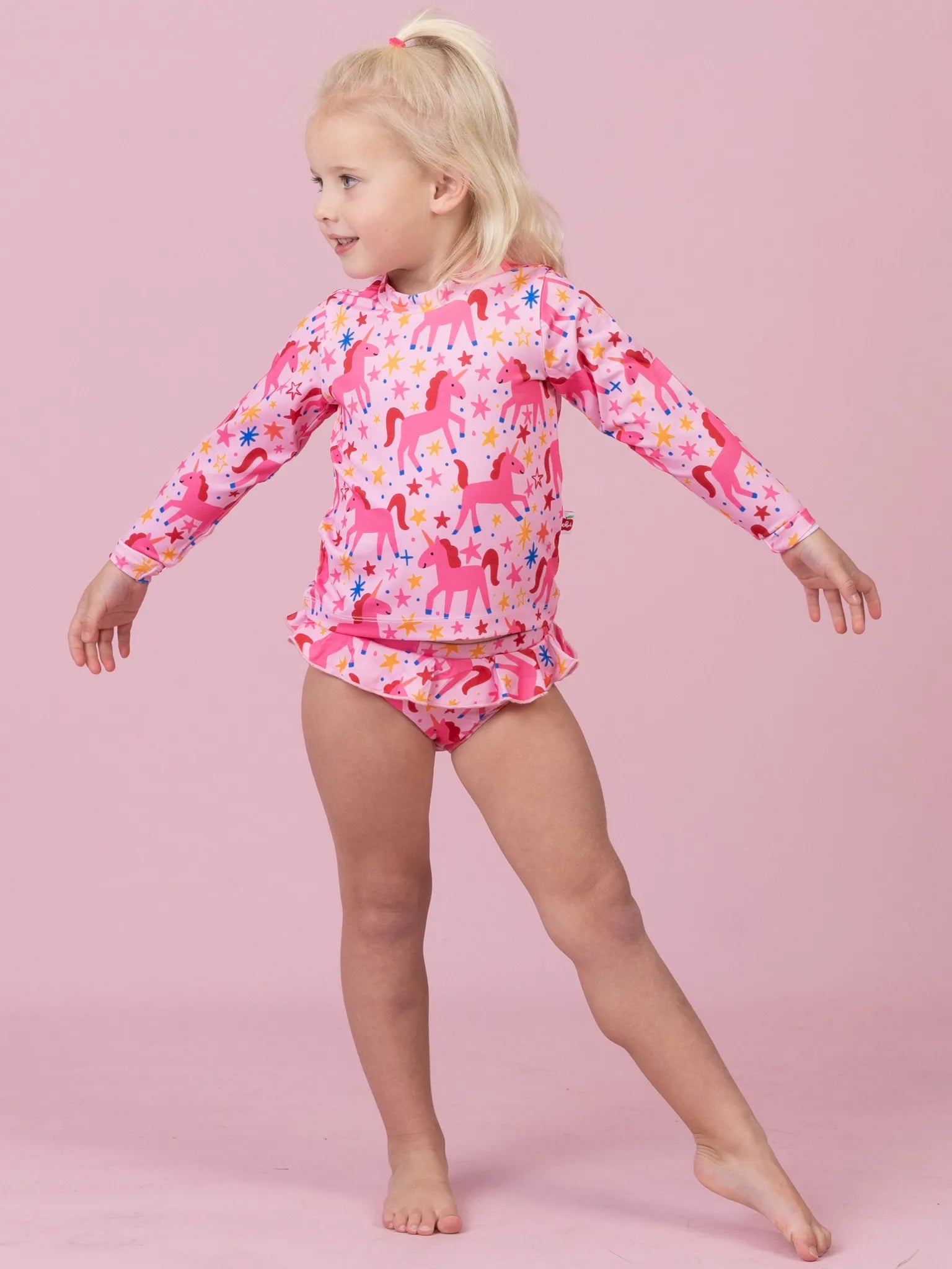 A young blonde girl modeling a pink long-sleeve swim shirt and matching swim briefs with a colorful unicorn print, posed against a solid pink background.