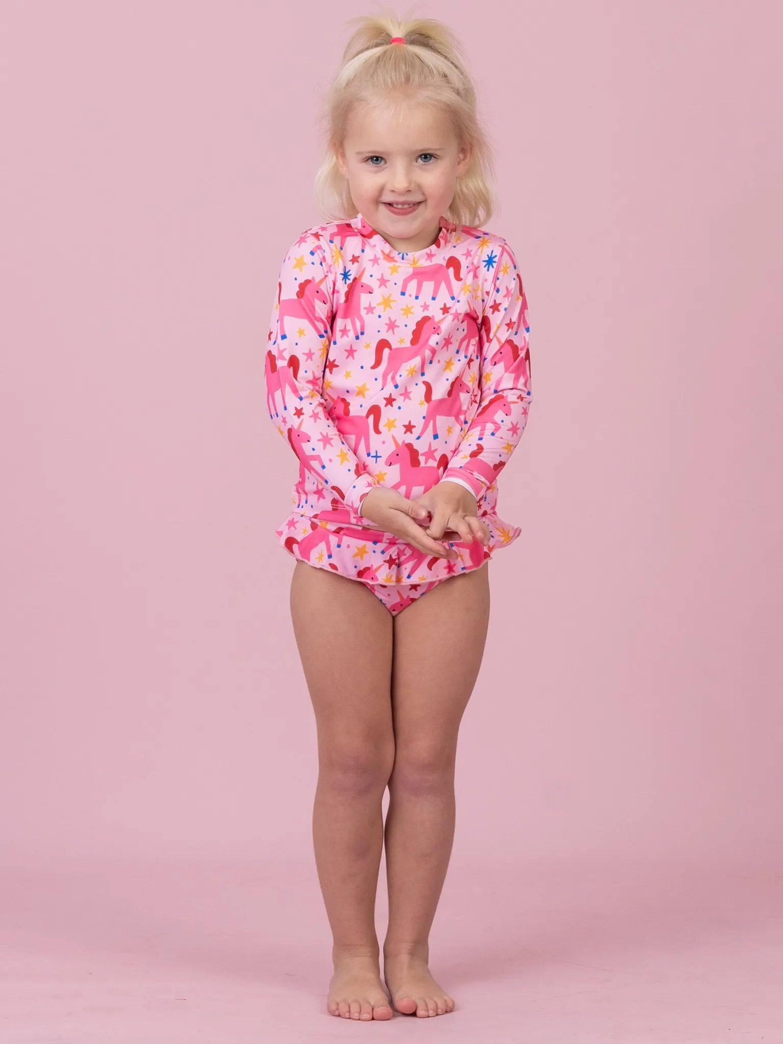 A young girl with blonde hair in a ponytail stands against a solid pink background, wearing a long-sleeve pink swimsuit with a colorful unicorn pattern.