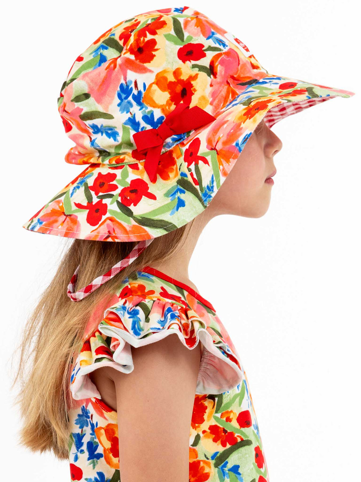 A child wearing a Wild Roses Chloe Ponytail Hat in vibrant floral colors, featuring a wide brim, along with a dress that has ruffled sleeves, is facing sideways against a white background.