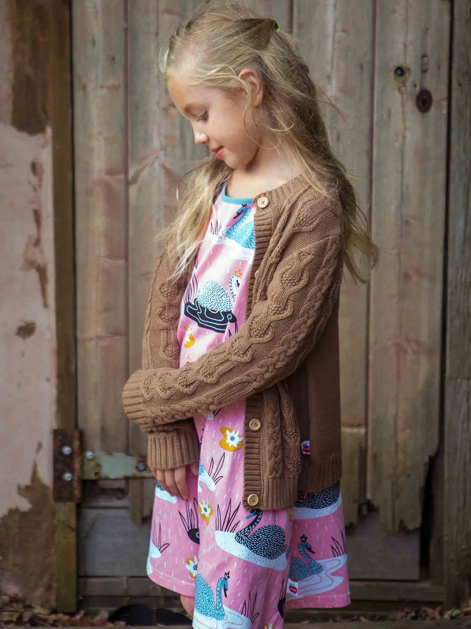 Young girl wearing a brown cardigan over a colorful dress, standing in front of a wooden door.