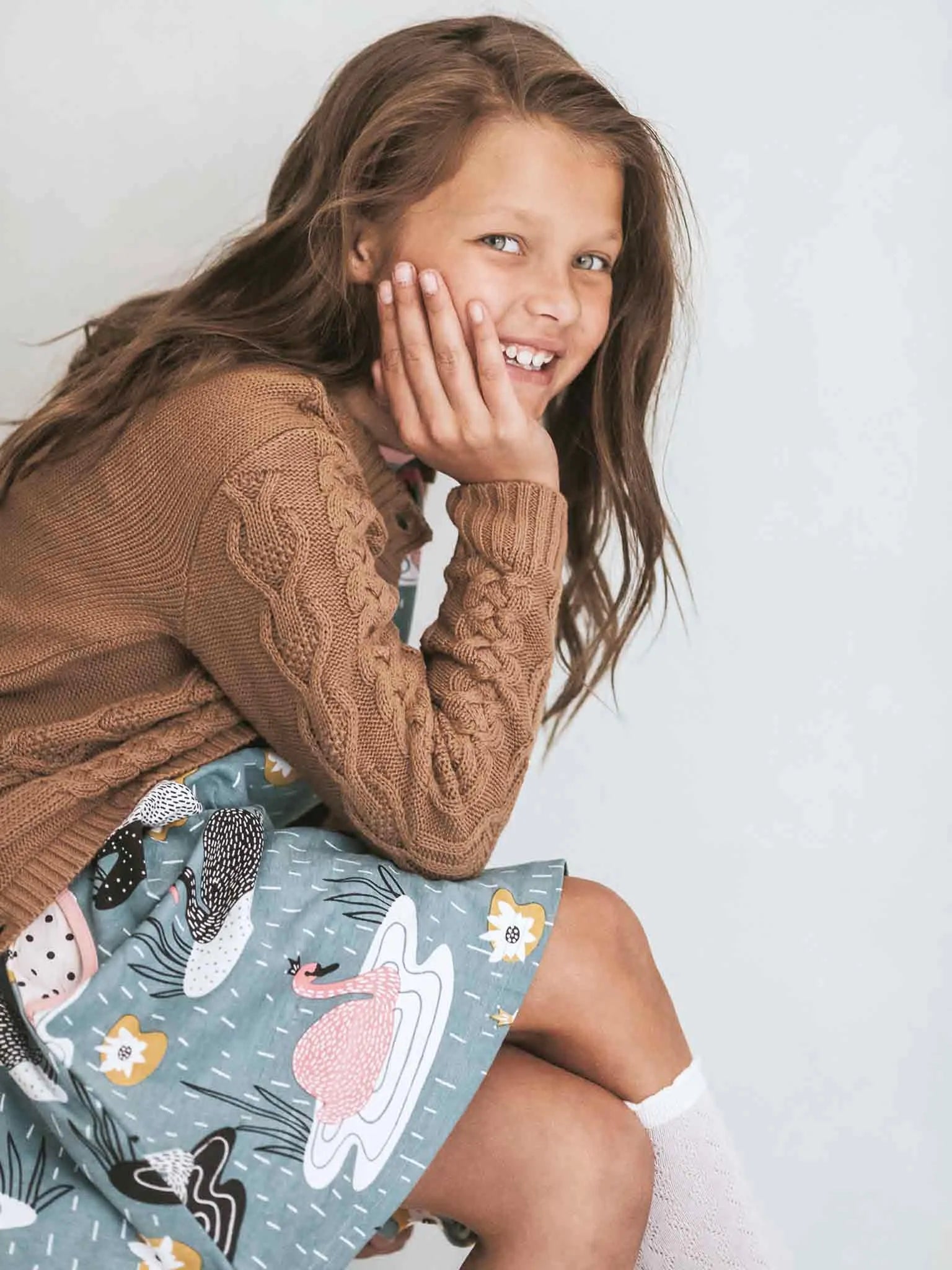 Young girl wearing a brown cable knit sweater and a patterned skirt on a white background