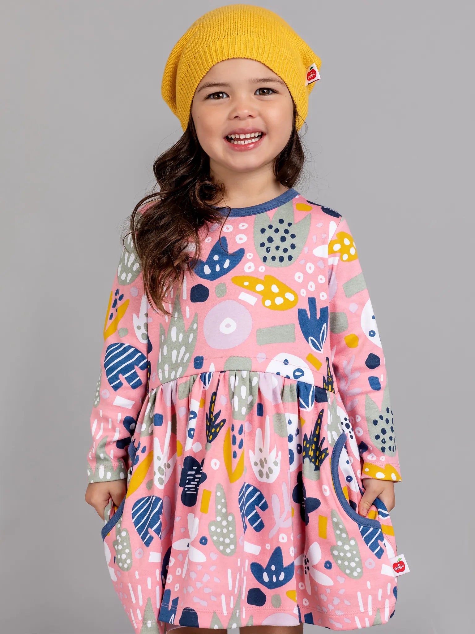 Child wearing a colorful dress with abstract patterns and a yellow beanie on a gray background
