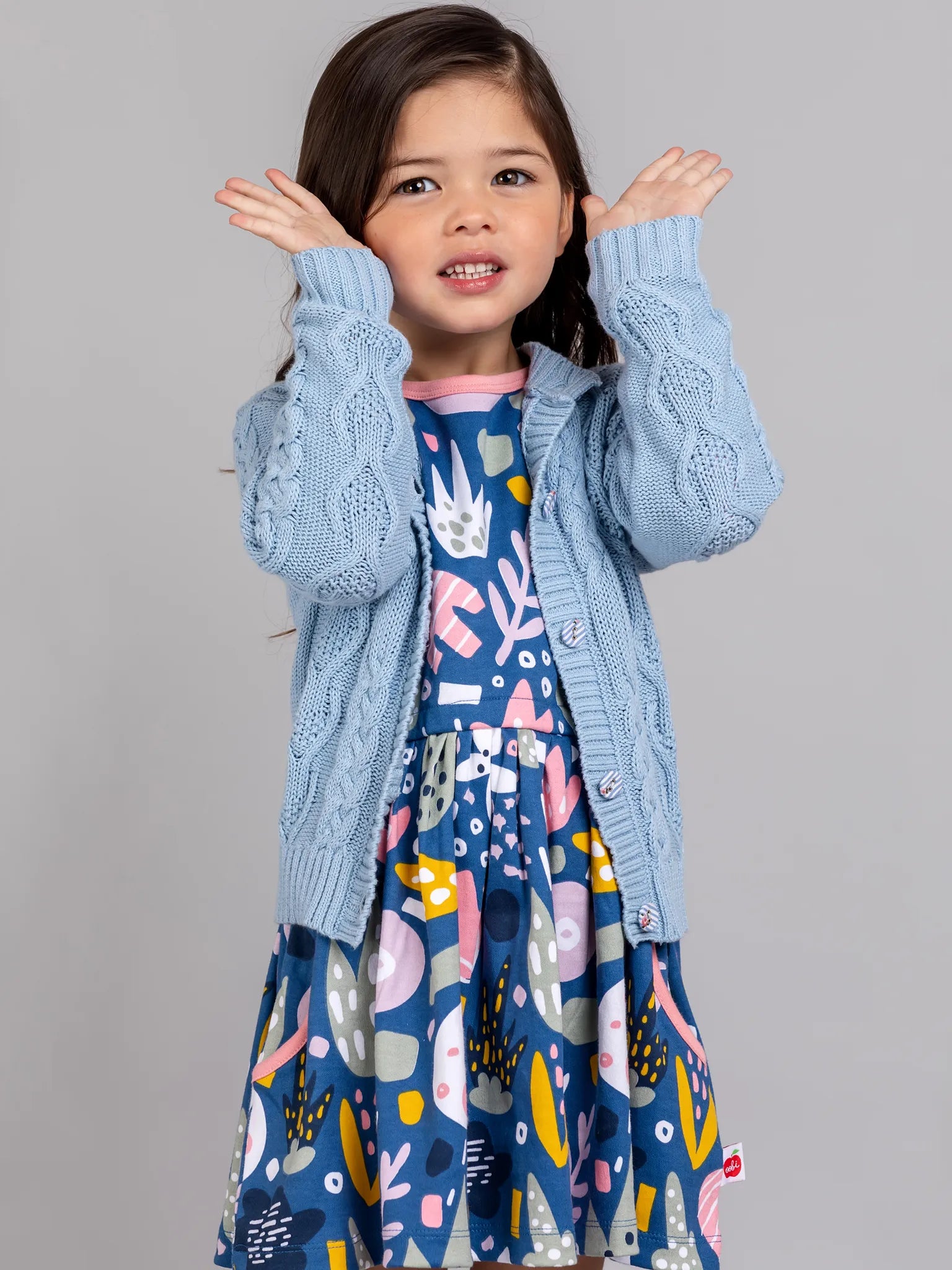 Young girl wearing a blue patterned dress with a light blue cardigan against a gray background