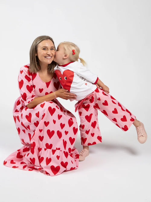 A woman and a young child wearing matching pink pajamas with a red heart pattern, posed together against a plain white studio background.