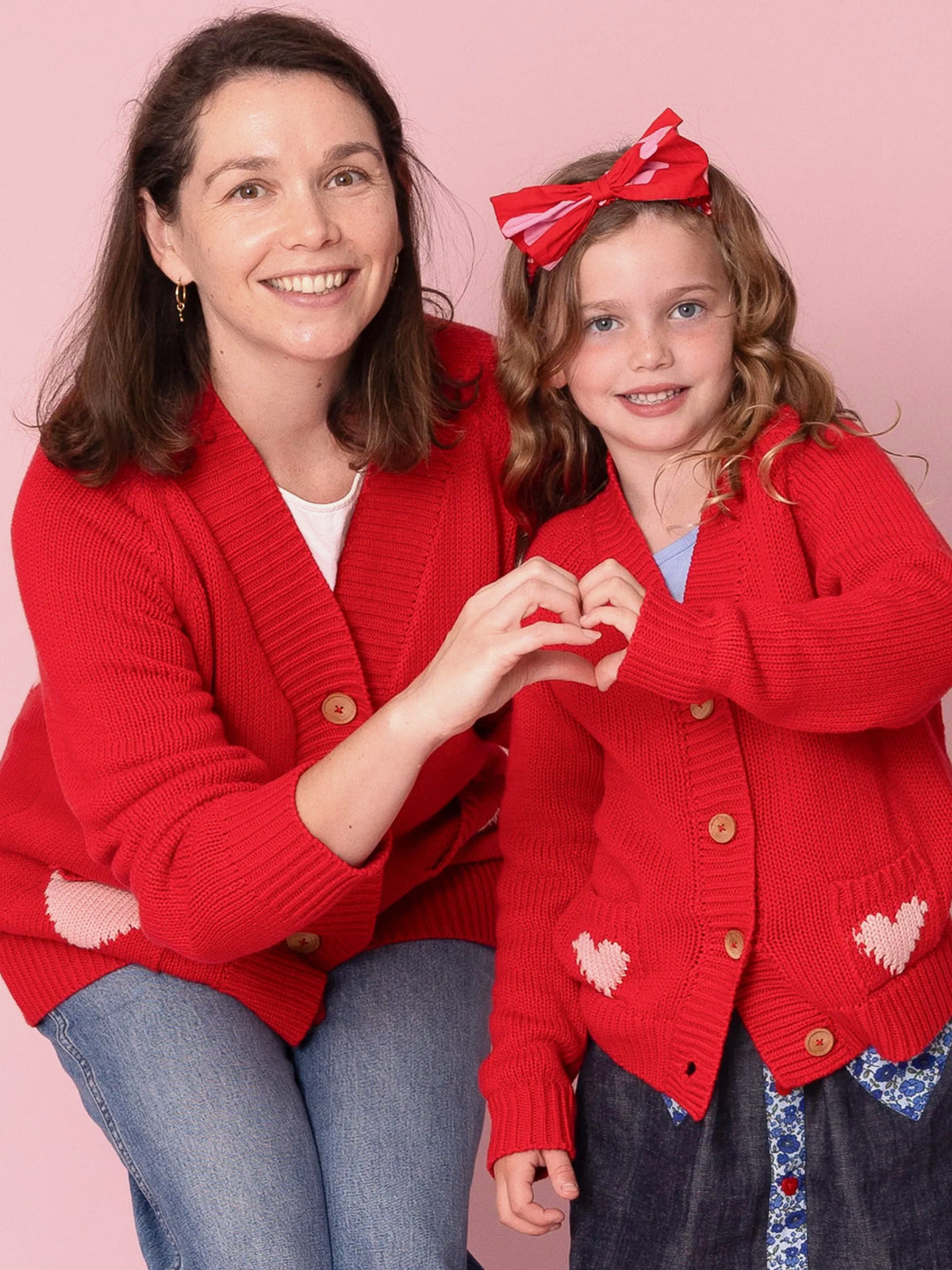 Woman and young girl wearing red cardigans with heart patterns against a pink background