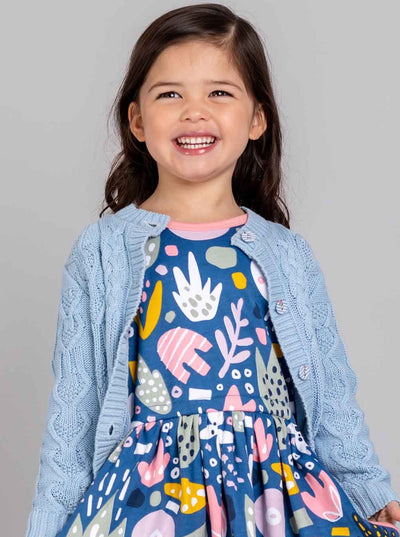 A smiling young girl wearing a patterned dress and a high-quality Knitwear Willow Sky Blue Cotton Cardigan.